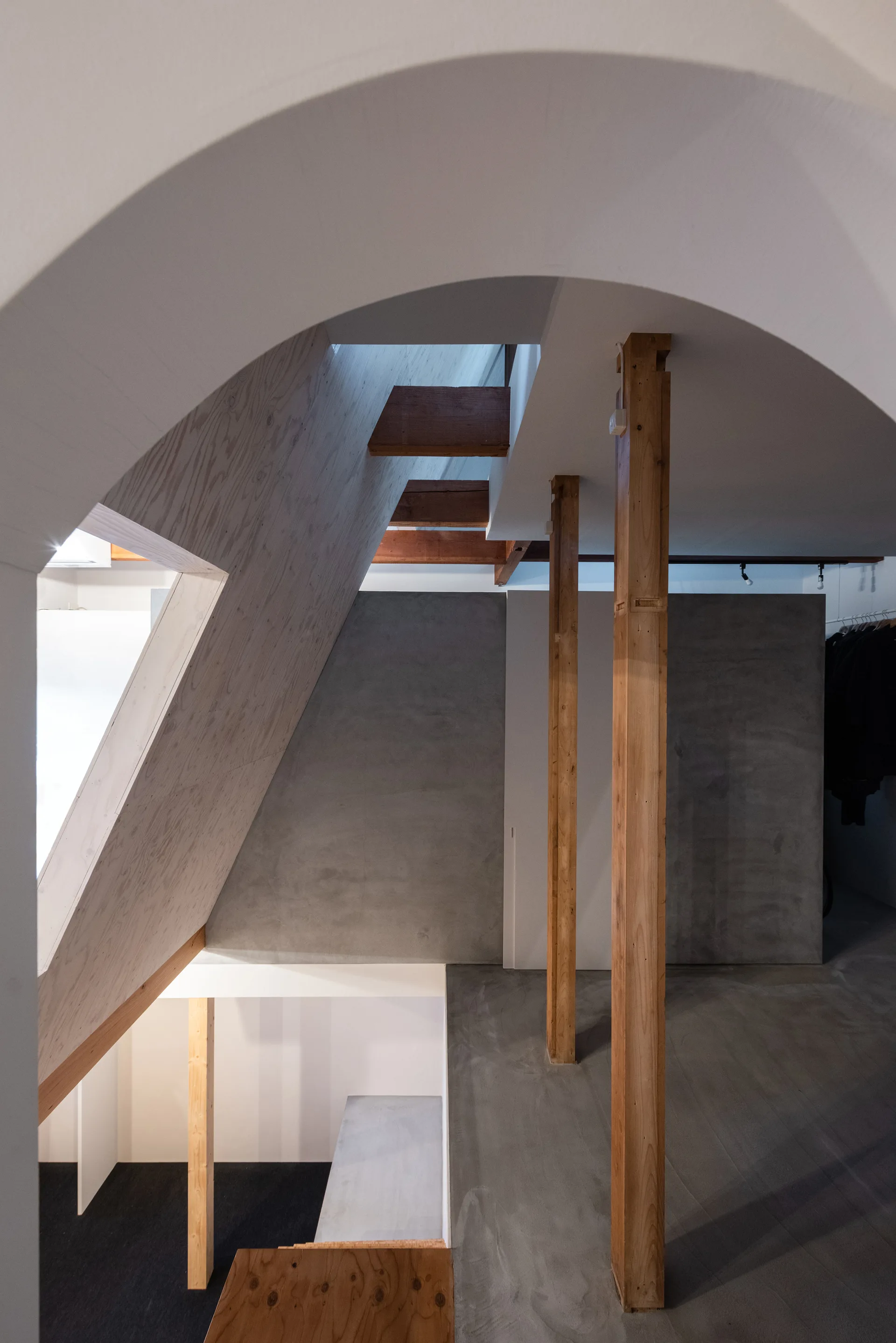 A modern minimalist stairwell features exposed wooden posts, plywood wall panels, concrete walls, and a large white plaster arch overhead.
