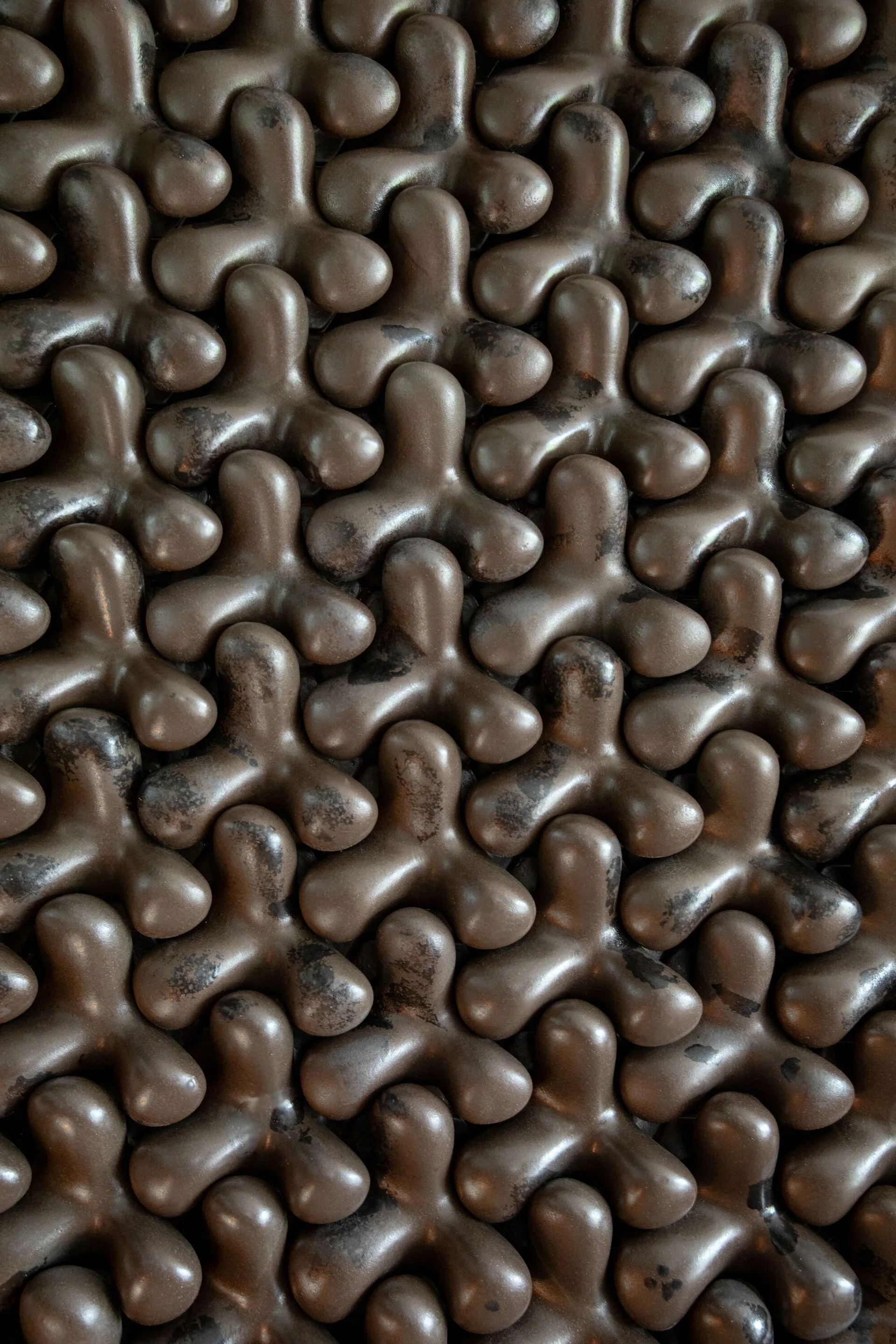 A close-up view of a wall covered in repeating, interlocking three-lobed dark bronze sculptural tiles with a patinated finish.