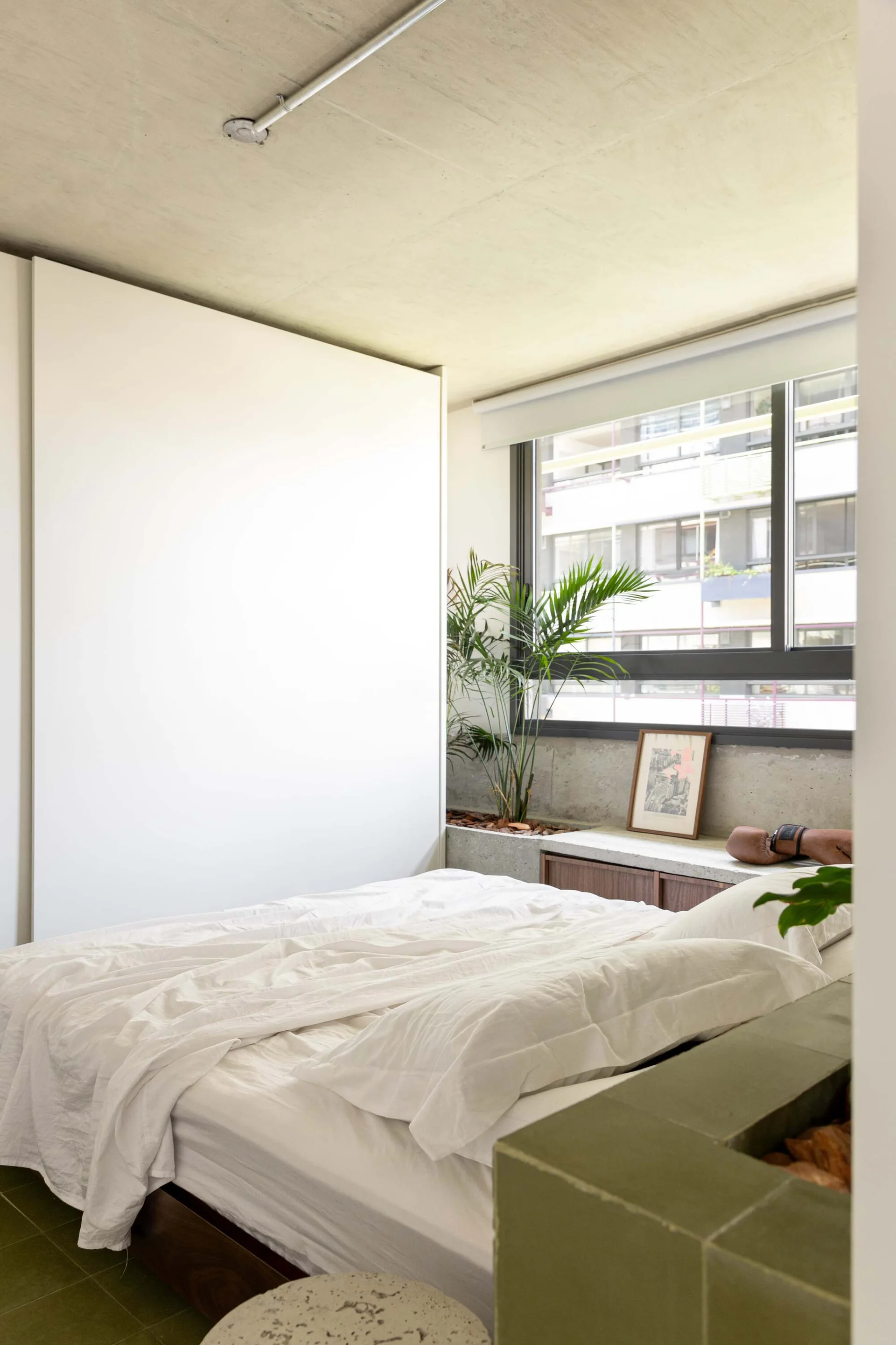A modern bedroom features white bedding, an exposed concrete ceiling, a large window, and a low built-in wood cabinet with a concrete top.