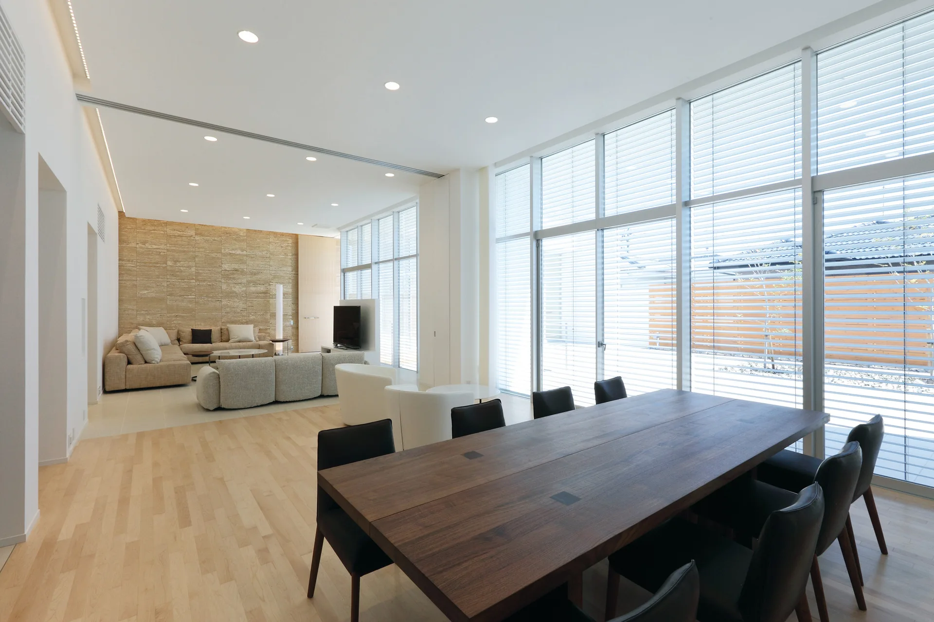 A minimalist open-concept space featuring a large wooden dining table with black chairs next to expansive windows with horizontal blinds.
