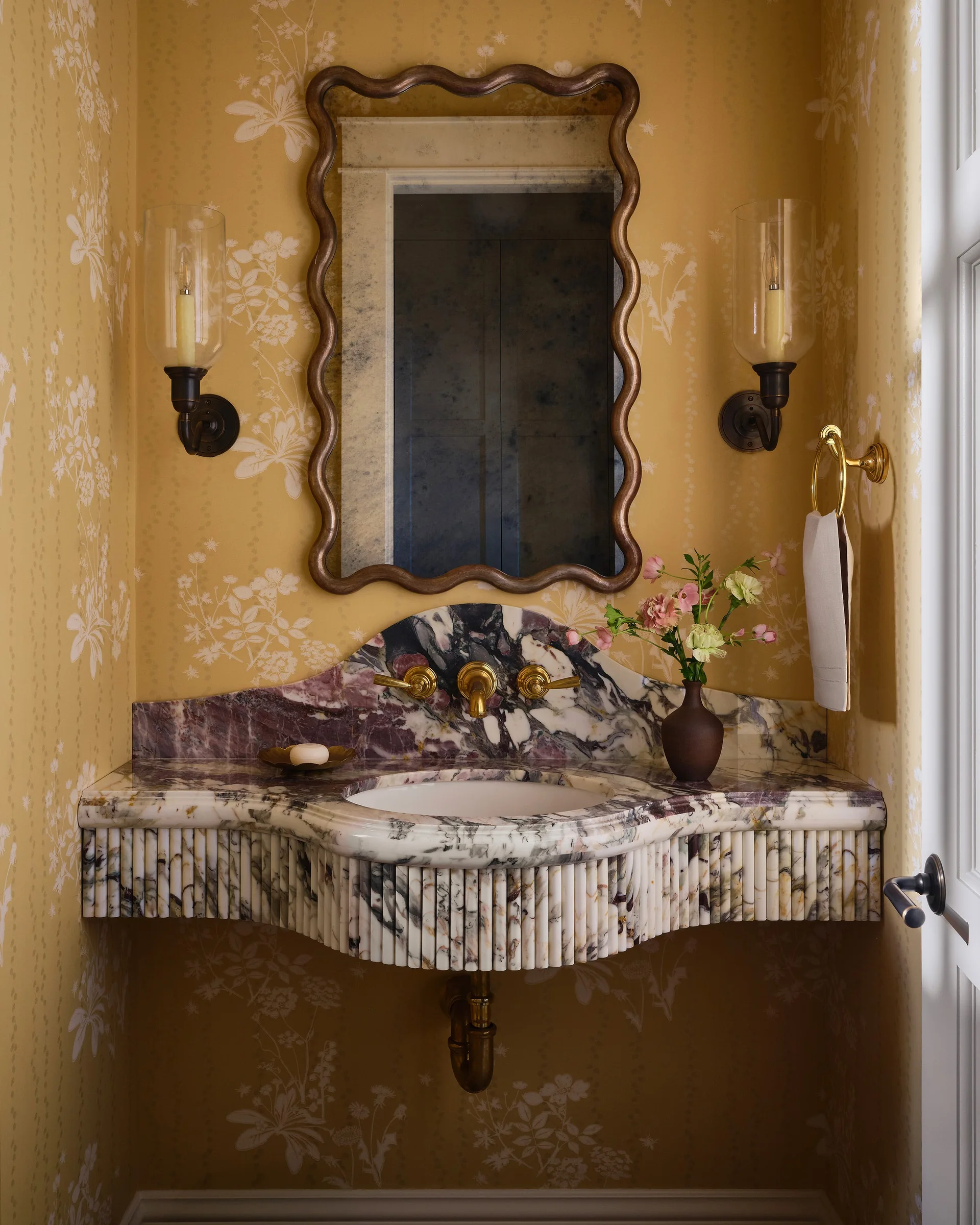 A powder room features a custom fluted marble vanity sink, gold wall-mounted faucet, scalloped mirror, and yellow floral wallpaper.