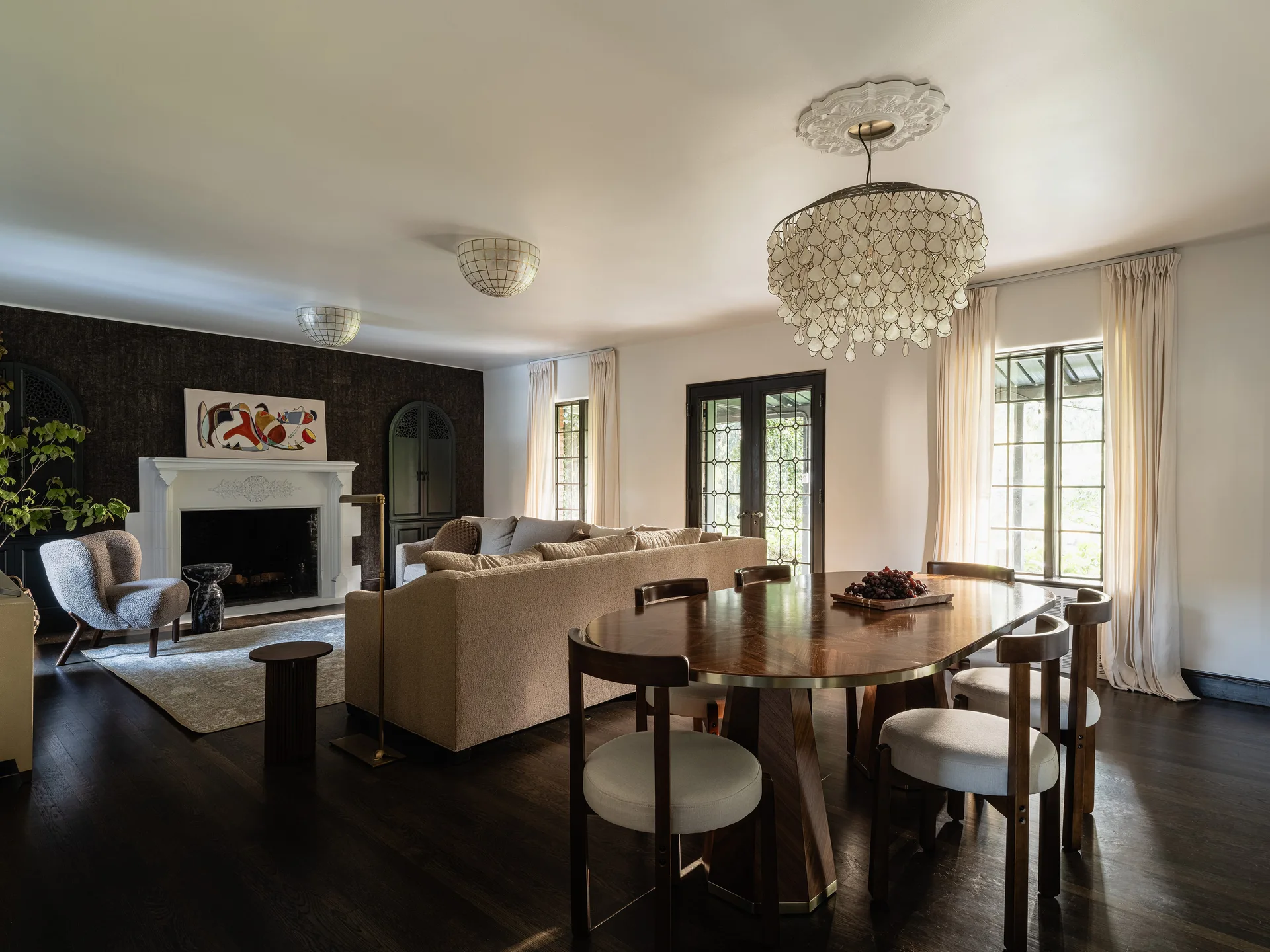An open-concept living and dining room with a dark wood table, neutral fabric sofa, and a large shell chandelier hanging from a ceiling medallion.