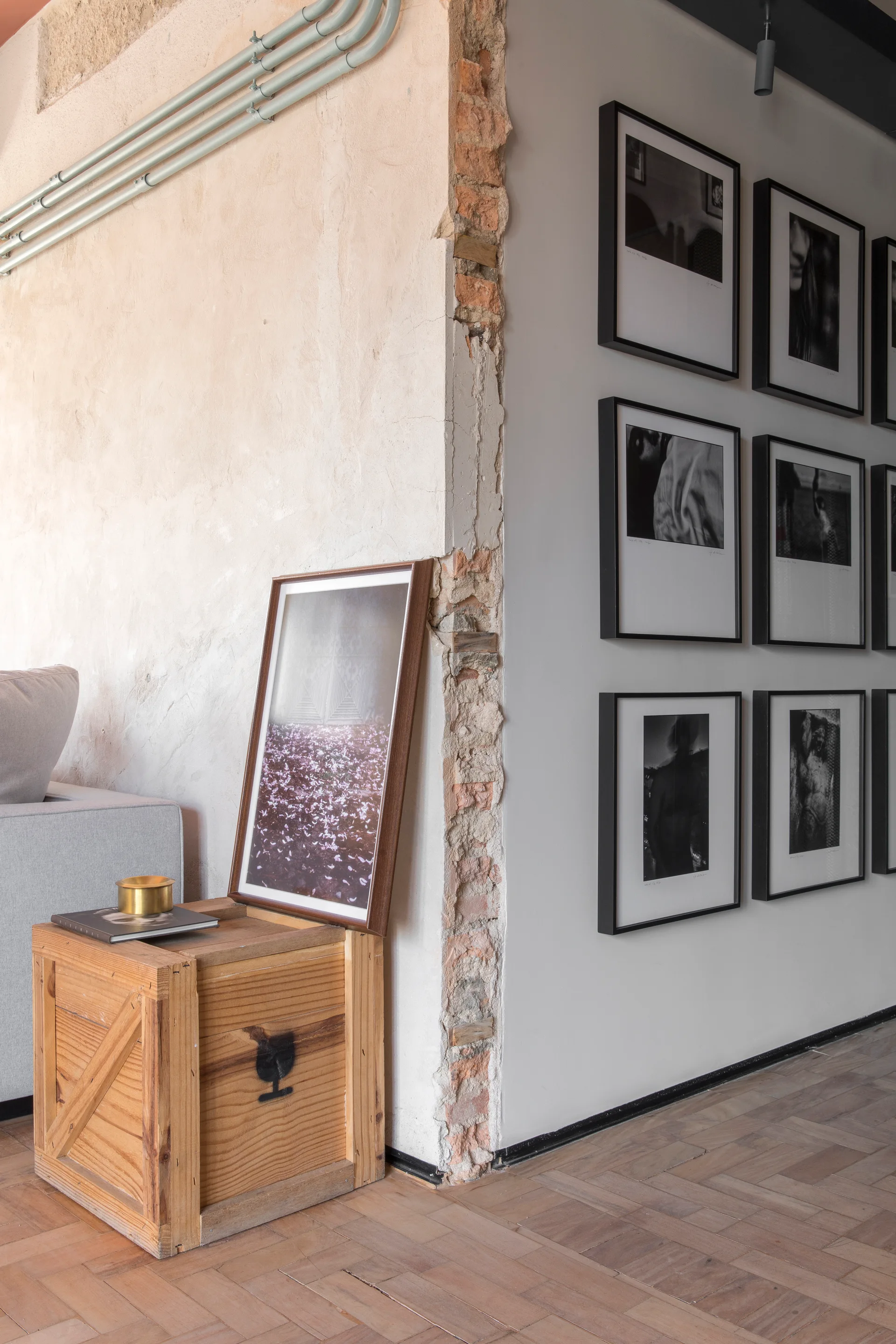 An interior space features a grid of black-framed monochrome photos on a wall, a corner with exposed brick, and a wooden crate side table.
