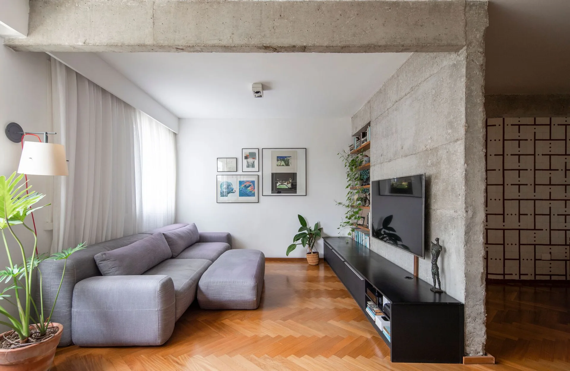 A contemporary living room featuring a grey sectional sofa on herringbone wood floors, a raw concrete wall, and a long black media console.