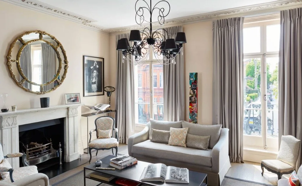 A cream living room featuring a grey sofa, a white fireplace with a large round gold mirror above, and an ornate black chandelier.