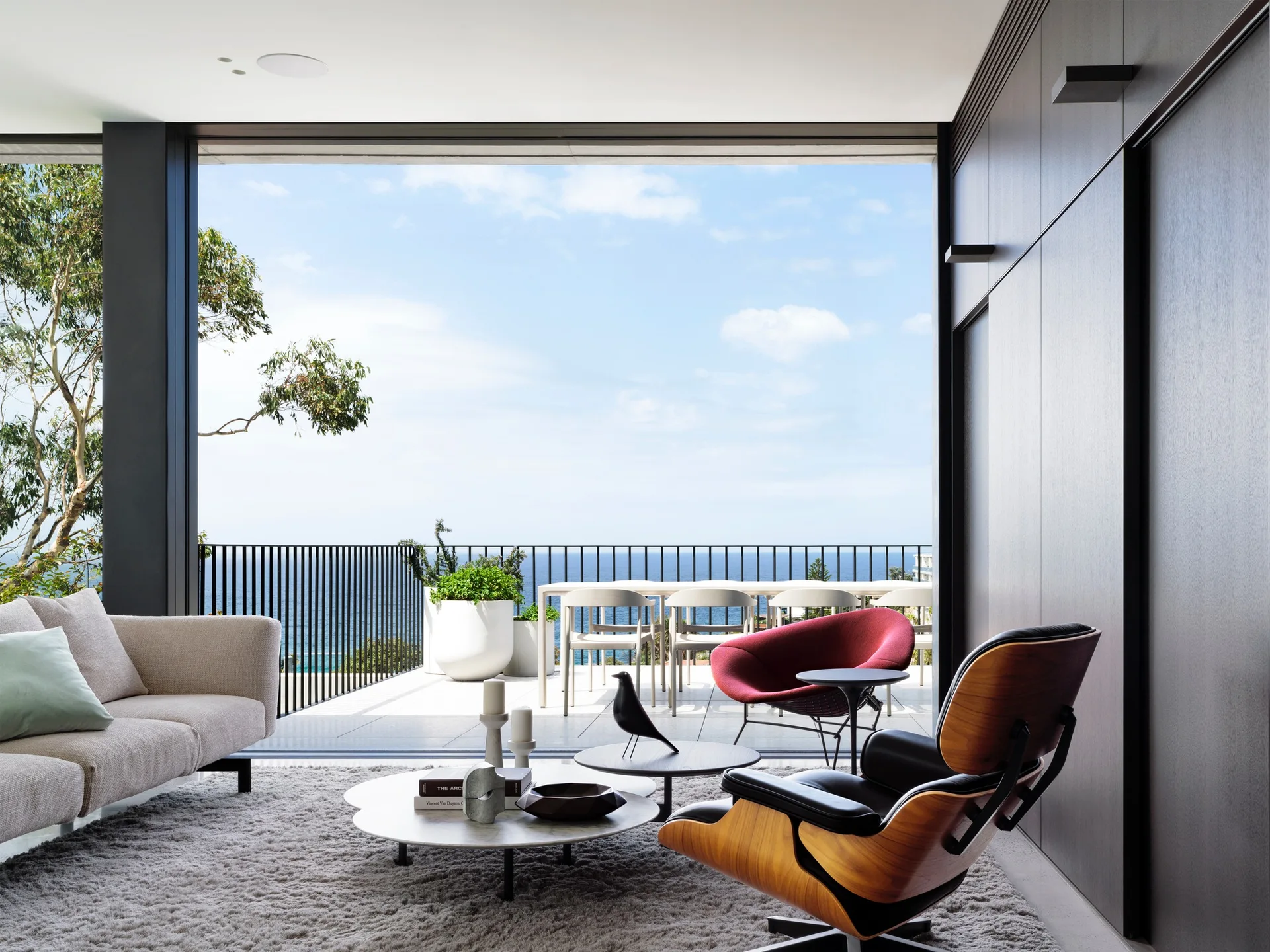 A modern living room with a leather lounge chair and cream sofa features a wide opening to an oceanfront terrace with outdoor seating.