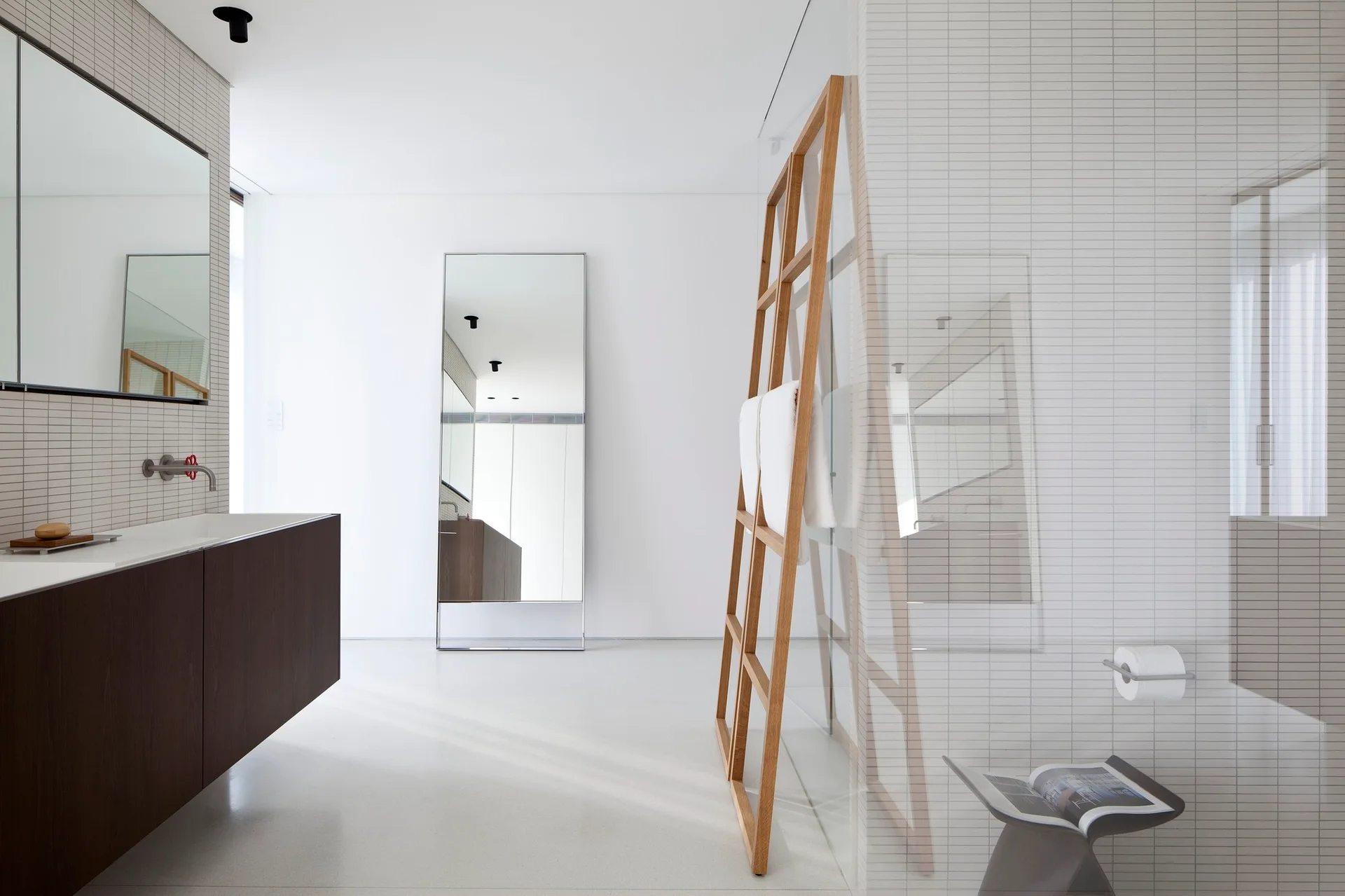 A minimalist white bathroom featuring a dark wood floating vanity, a tall floor mirror, and a wooden ladder towel rack next to a glass shower panel.