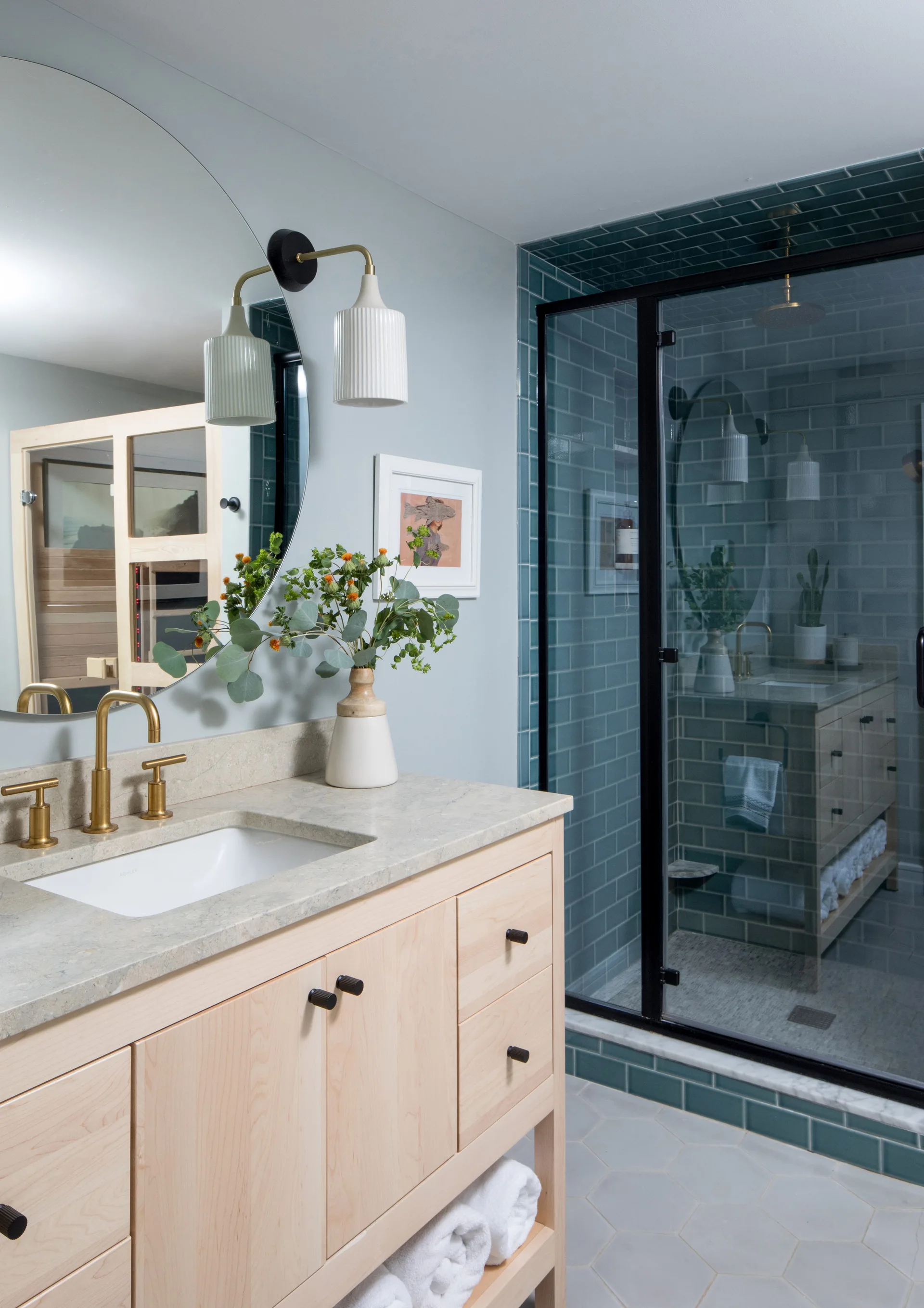 A modern bathroom features a light wood vanity with a stone countertop, a round mirror, and a glass shower enclosure with teal subway tiles.