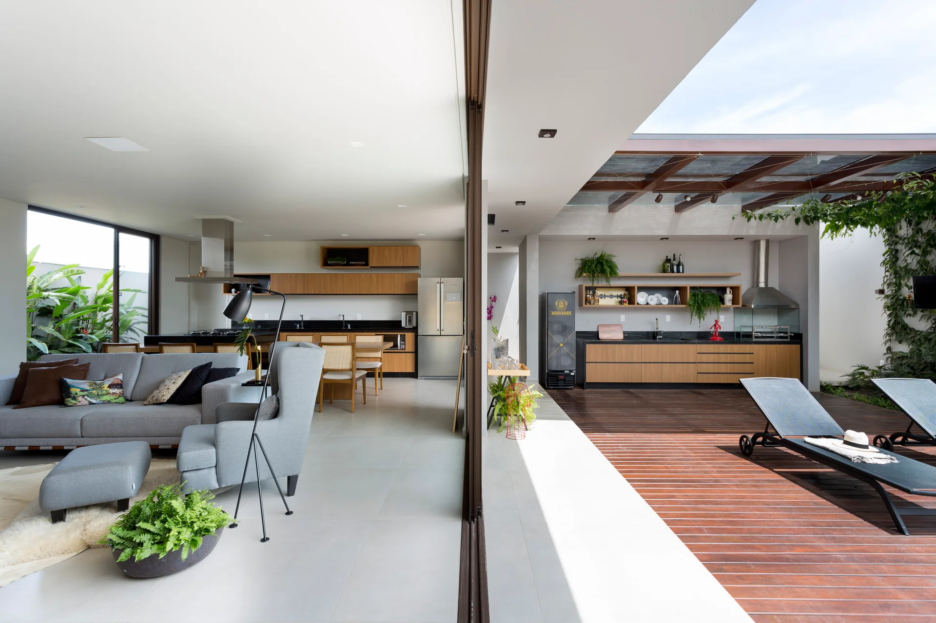 A sliding glass partition connects an indoor grey living room and kitchen to an outdoor wooden deck with a built-in patio kitchen.
