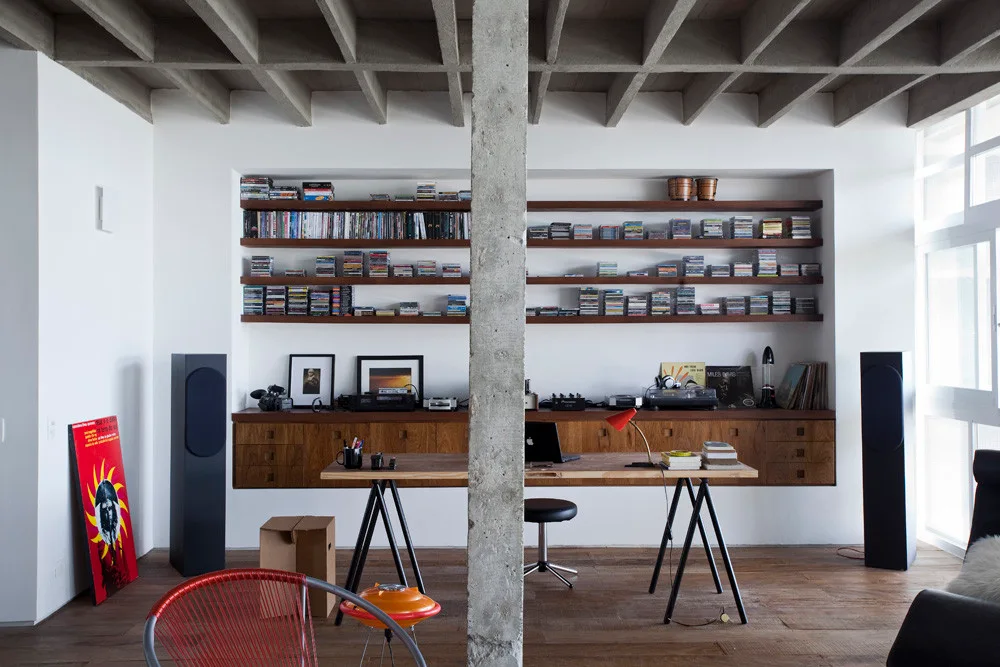 A minimalist home office featuring a wooden trestle desk, recessed shelving filled with media, and a prominent concrete ceiling and column.