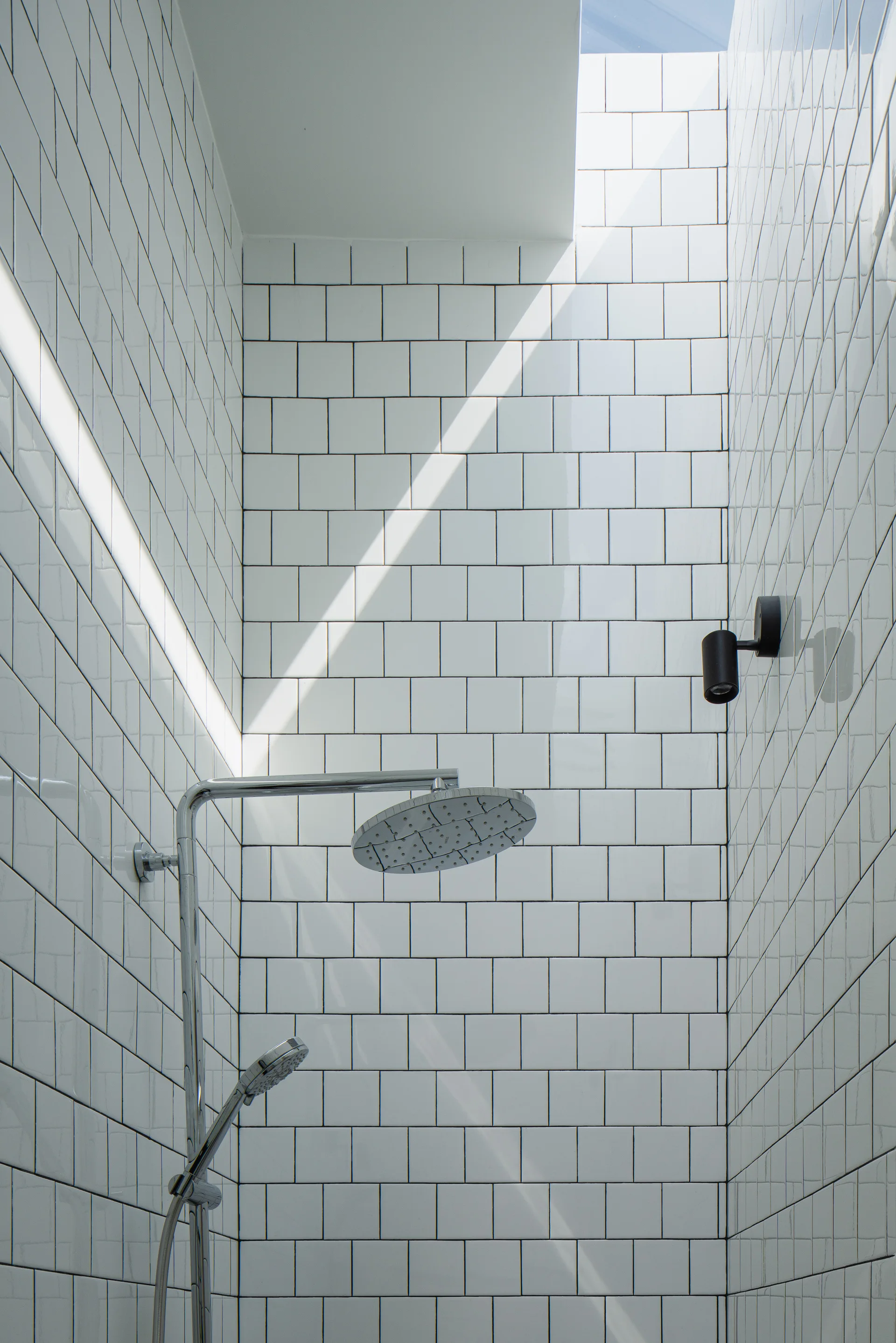 A modern shower features glossy white square wall tiles with dark grout, a chrome shower head assembly, a black sconce, and an overhead skylight.