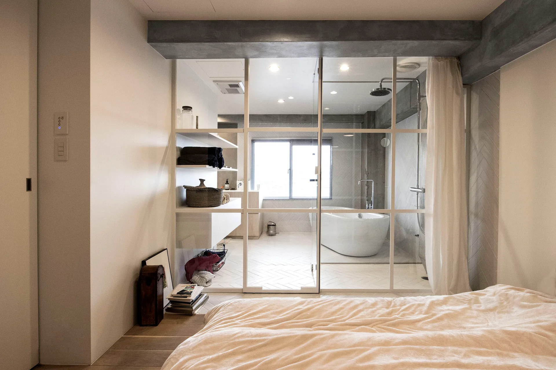A bedroom view looking through a white grid glass partition into an ensuite bathroom with a freestanding tub and a concrete ceiling beam.