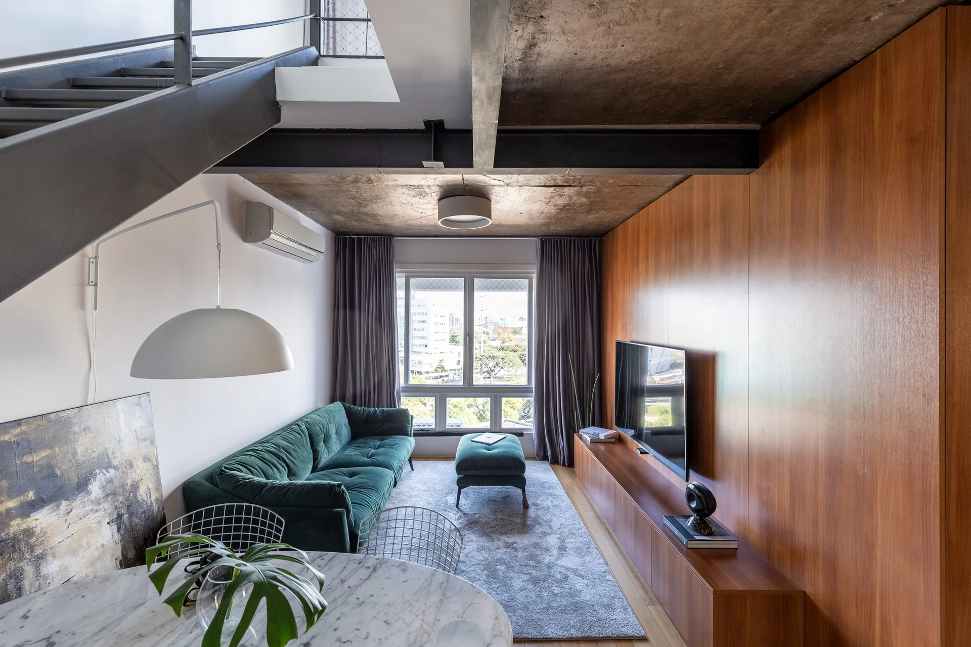 A modern loft living room features a dark green tufted sofa, a wood-paneled wall with a mounted TV, and an exposed concrete ceiling under a metal staircase.