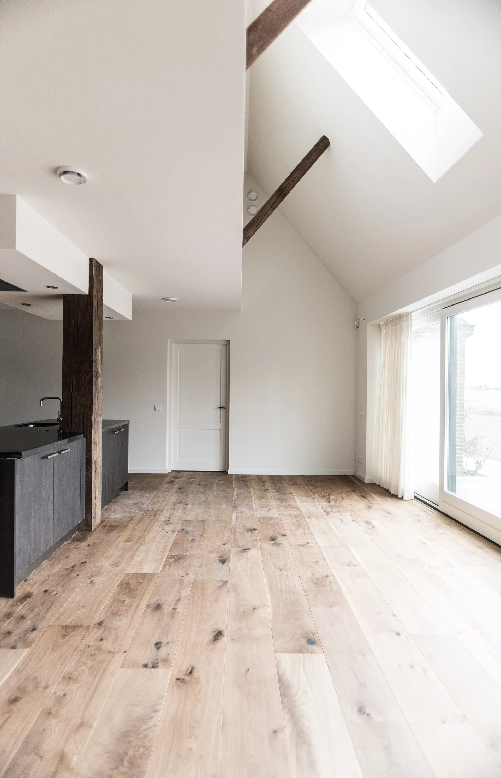 An empty open-plan kitchen with wide plank wood floors, white vaulted ceilings with exposed wood beams, and charcoal grey cabinetry.