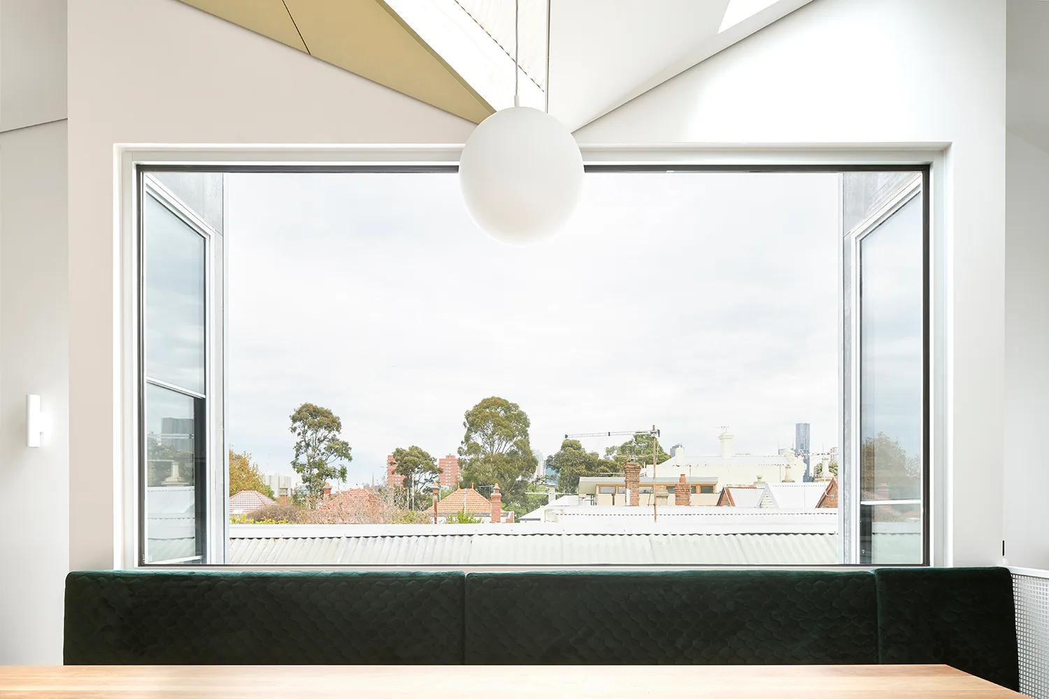 A dark green quilted bench sits below a large rectangular window framing a neighborhood view, with a white sphere pendant light hanging above.