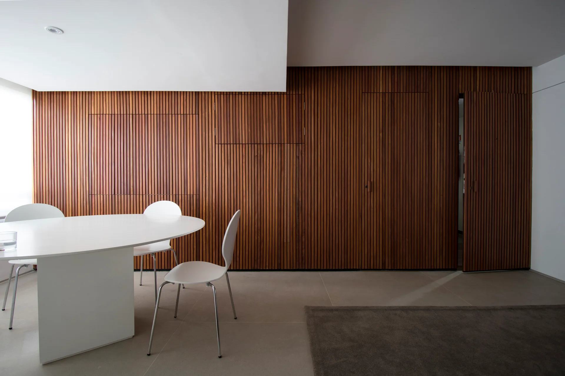 A white oval dining table and matching chairs stand in front of a floor-to-ceiling vertical slatted wood wall with integrated flush-mount doors.