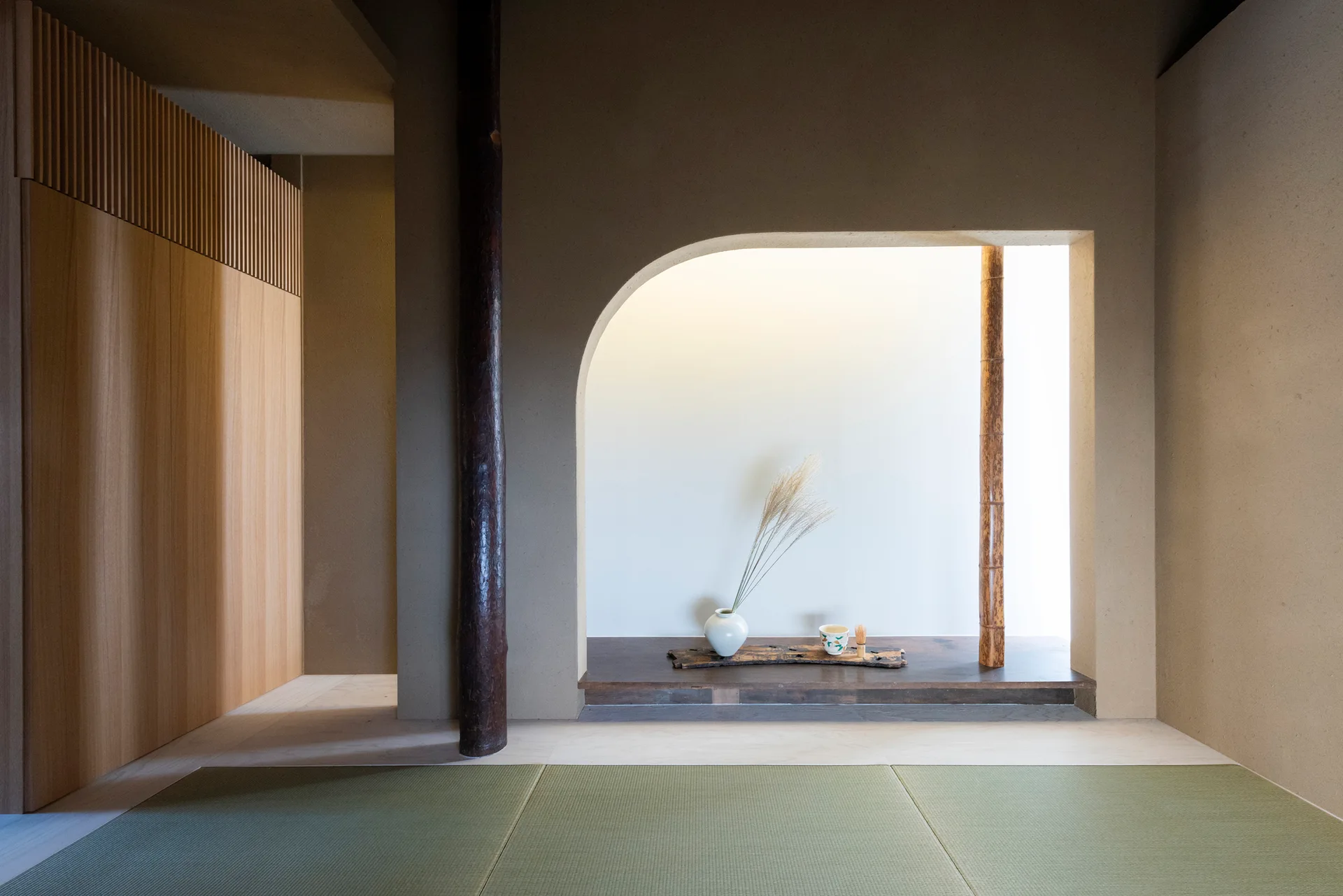 A minimalist Japanese room features green tatami mat flooring, a rustic wood pillar, and an arched tokonoma display alcove with a ceramic vase.