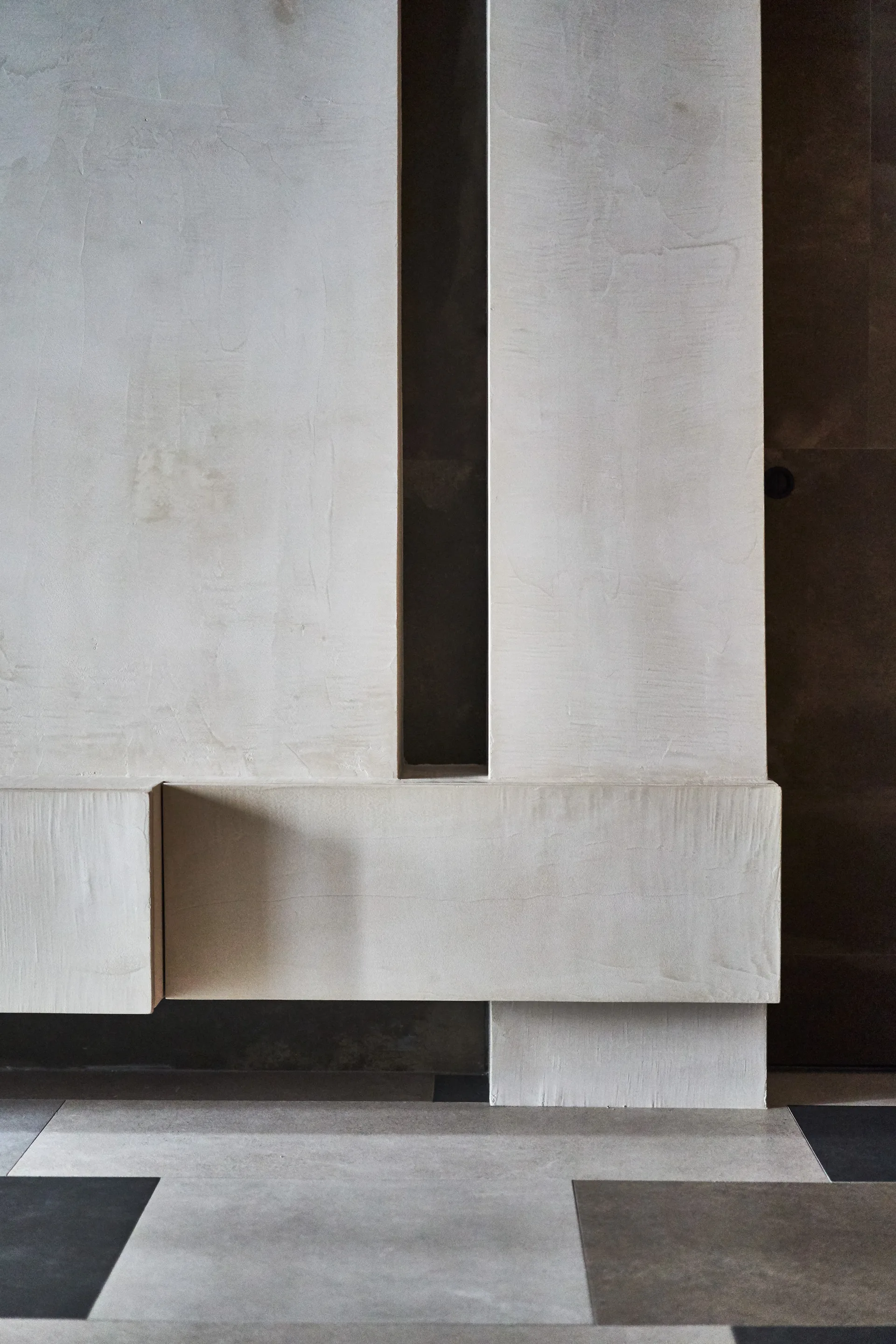 Layered cream-colored plaster wall panels feature a deep vertical shadow gap above a grey and charcoal large-format tiled floor.