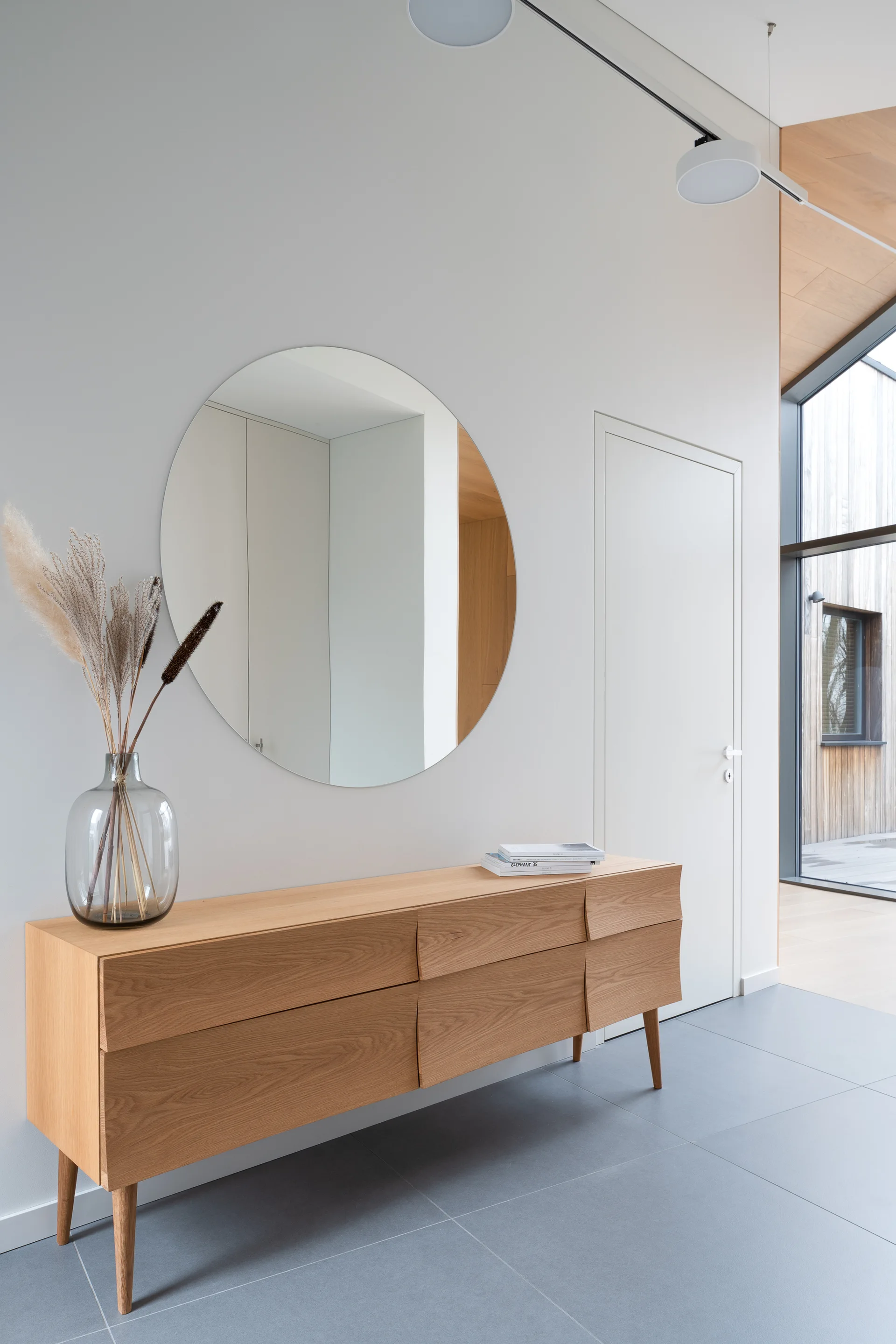 A minimalist entryway featuring a light oak console table, a large circular wall mirror, and a gray tiled floor.