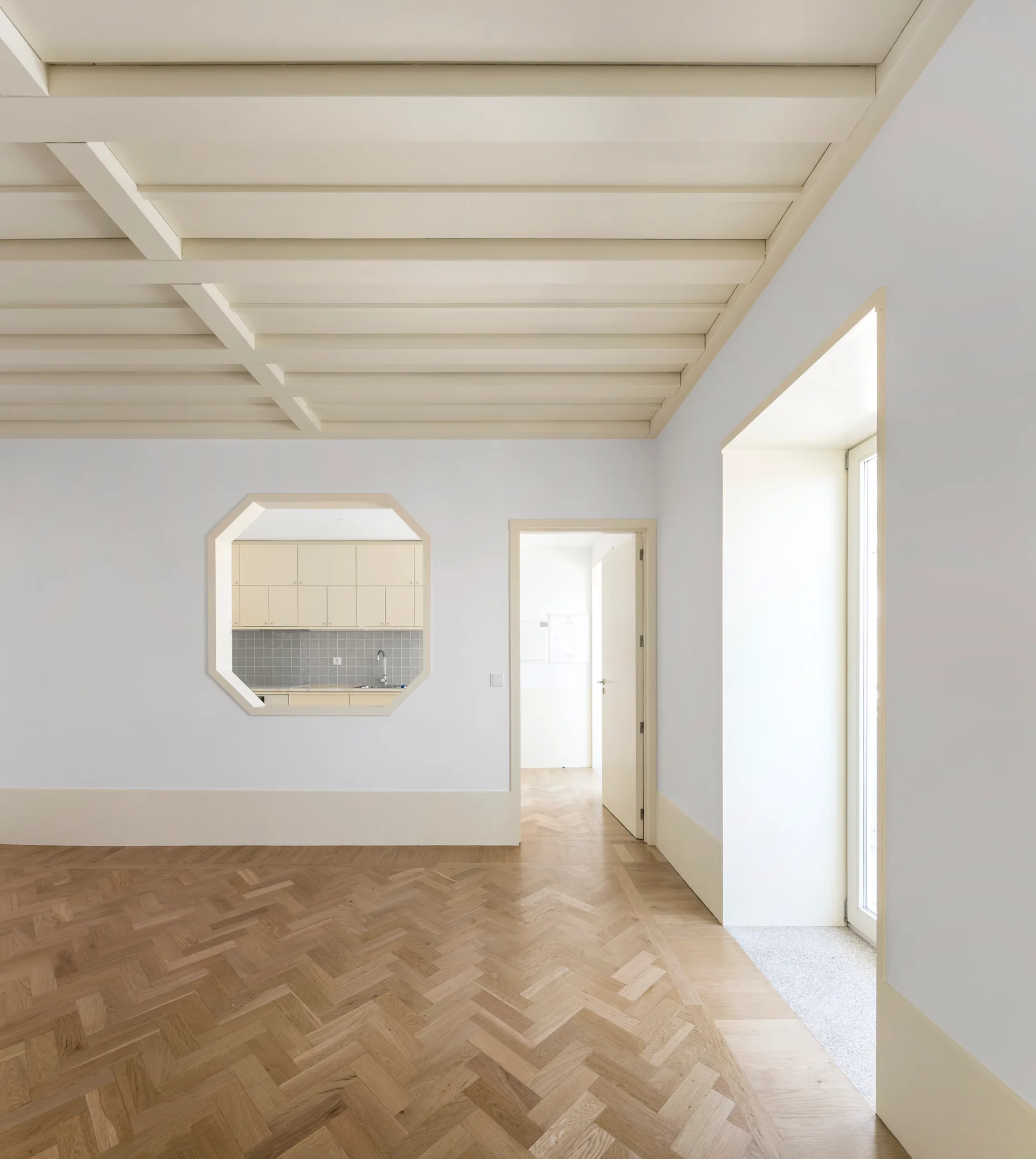 An empty room featuring light herringbone wood flooring, a painted beamed ceiling, and an octagonal cutout looking into a beige kitchen.