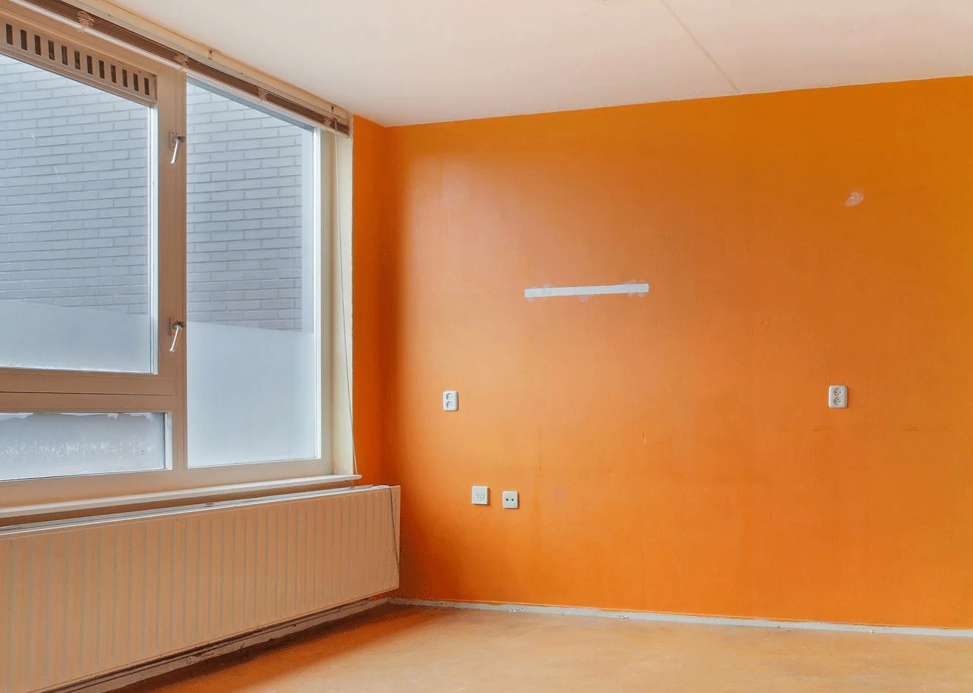 An empty interior room features a bold orange wall next to a large window and a white radiator.