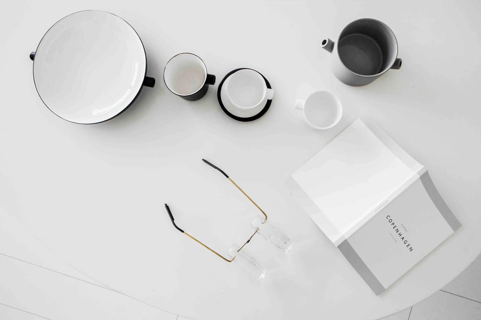 An overhead flat lay of minimalist black and white ceramic tableware, an open book, and glasses on a clean white tabletop.