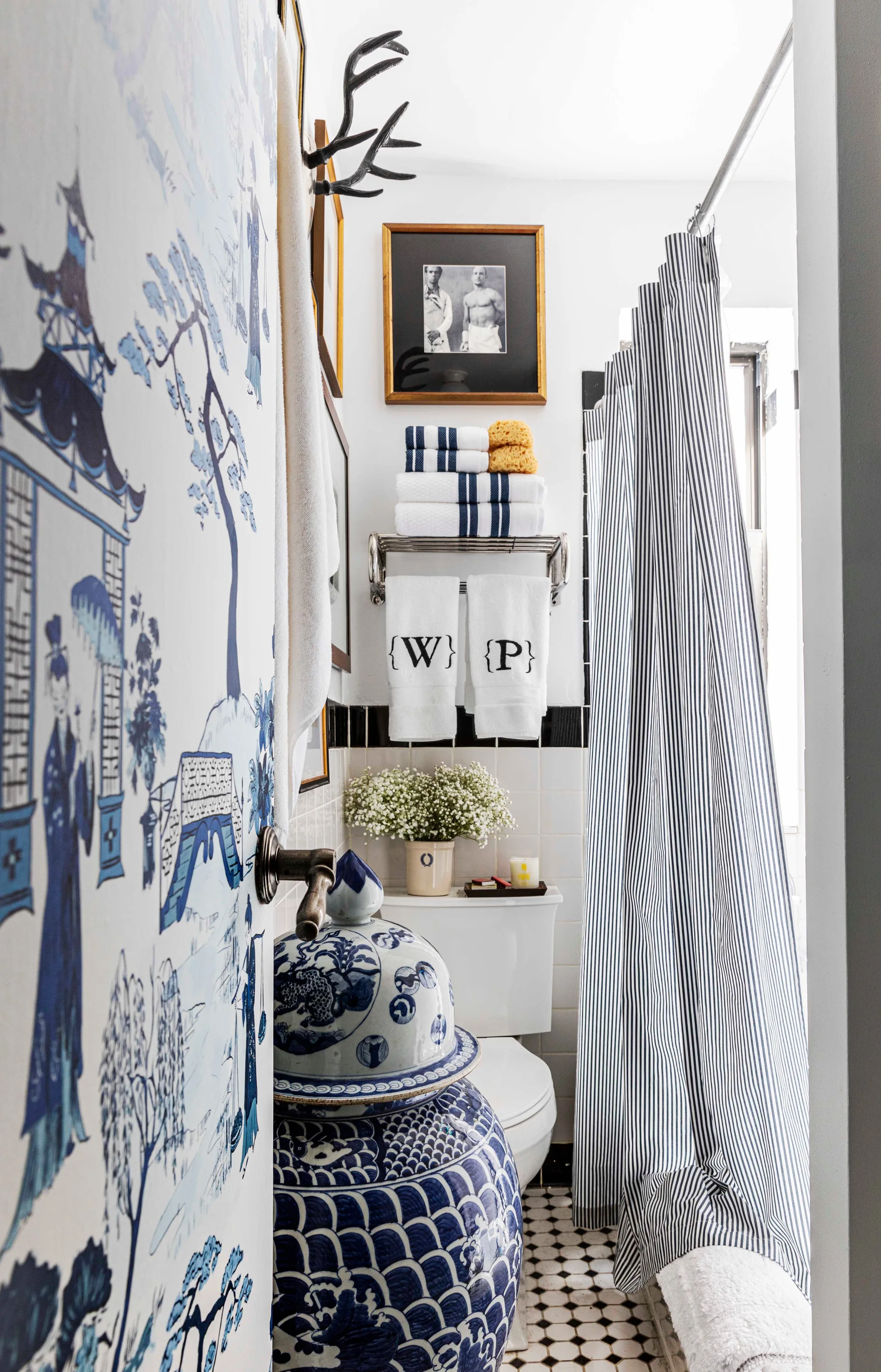 A small bathroom features blue and white chinoiserie wallpaper, a striped shower curtain, a white toilet, and monogrammed hanging towels.