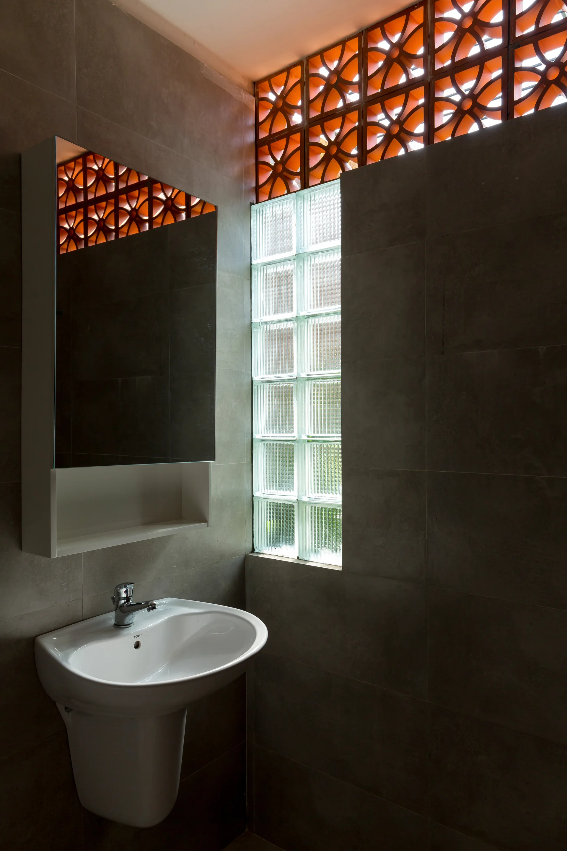 A white sink and mirrored cabinet in a gray-tiled bathroom featuring a glass block column and orange terracotta breeze blocks near the ceiling.