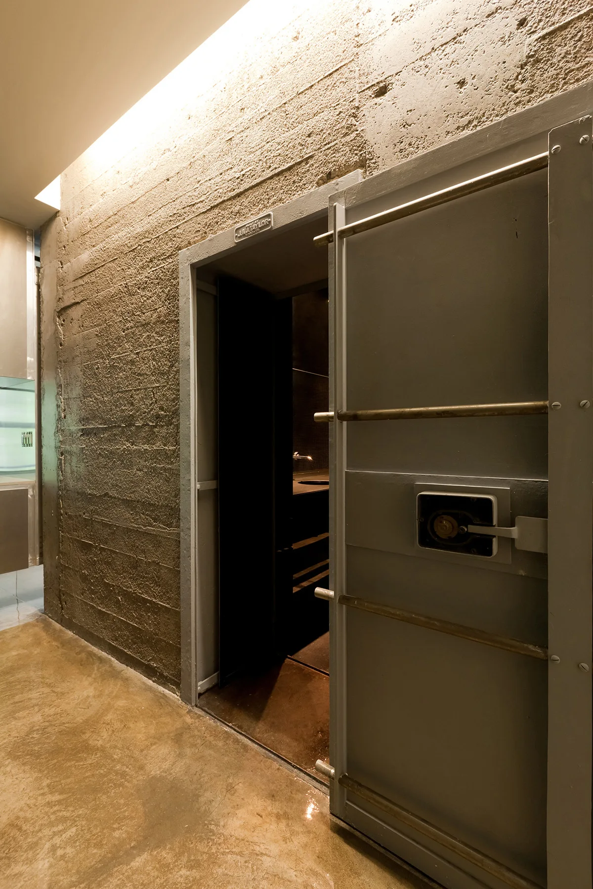 An open steel vault door installed on a textured, board-formed concrete wall with polished concrete floors and cove lighting.