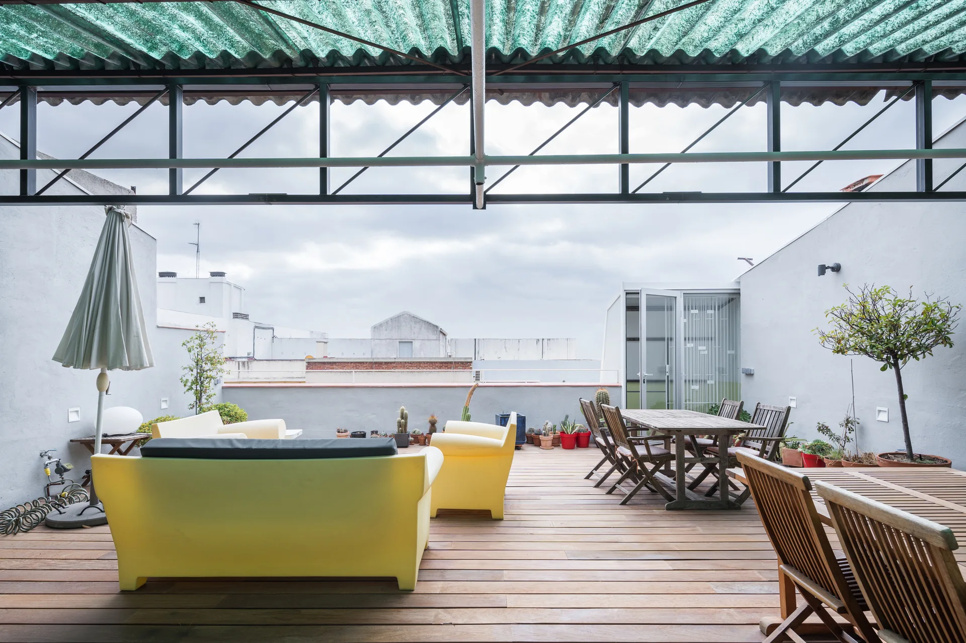 A spacious rooftop terrace features natural wood decking, bright yellow lounge chairs, a wooden dining table, and a green corrugated overhead awning.