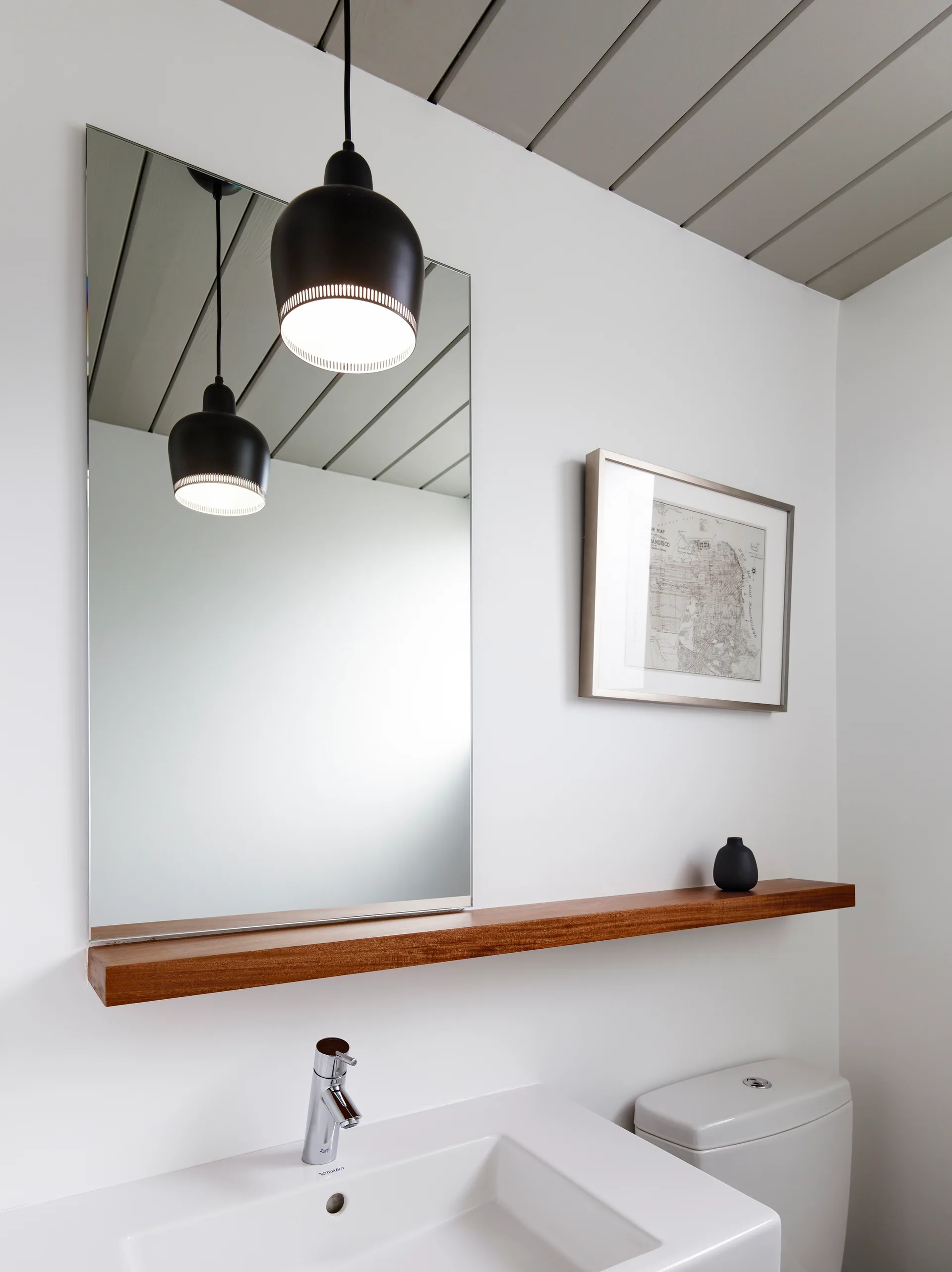 A minimalist bathroom features a white sink, a rectangular mirror reflecting a black pendant light, and a long floating wood shelf.