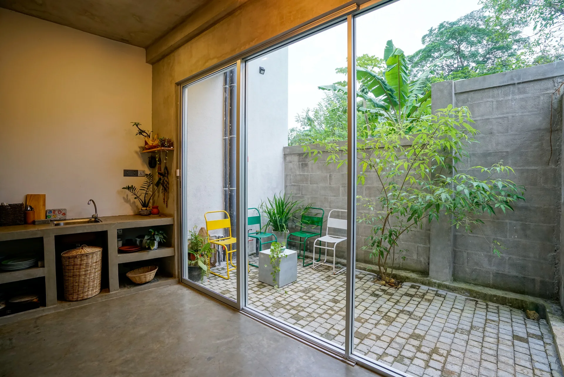 An industrial concrete kitchen counter sits next to a large sliding glass door that opens to a small paved patio with colorful metal chairs.