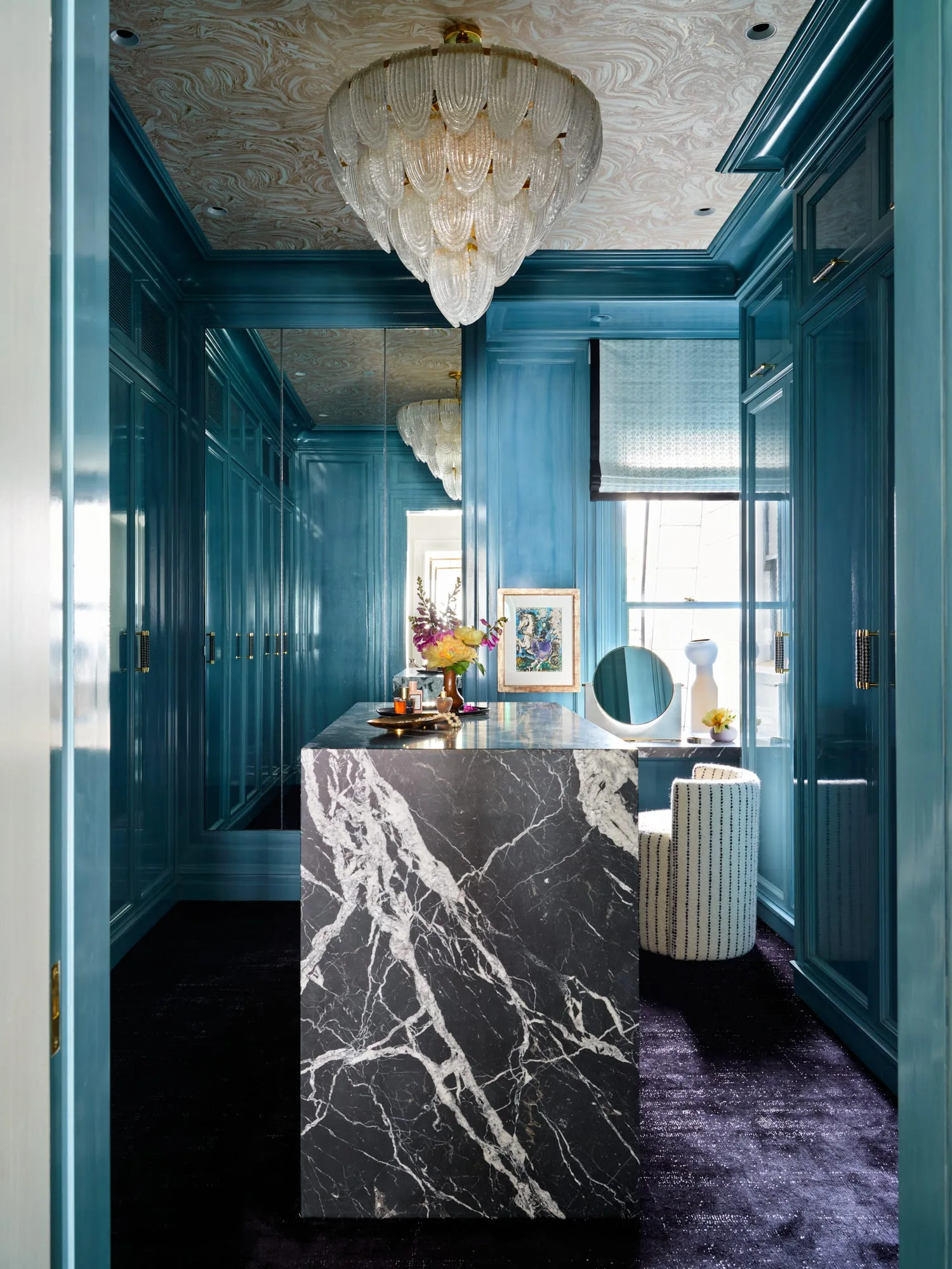 A dressing room features high-gloss teal cabinetry, a black and white veined marble island, and a tiered glass chandelier.