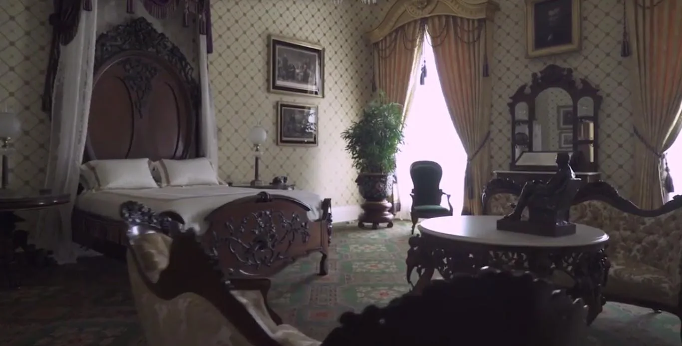 A Victorian bedroom featuring an ornate carved dark wood canopy bed, diamond-patterned wallpaper, and heavy drapes framing a window.