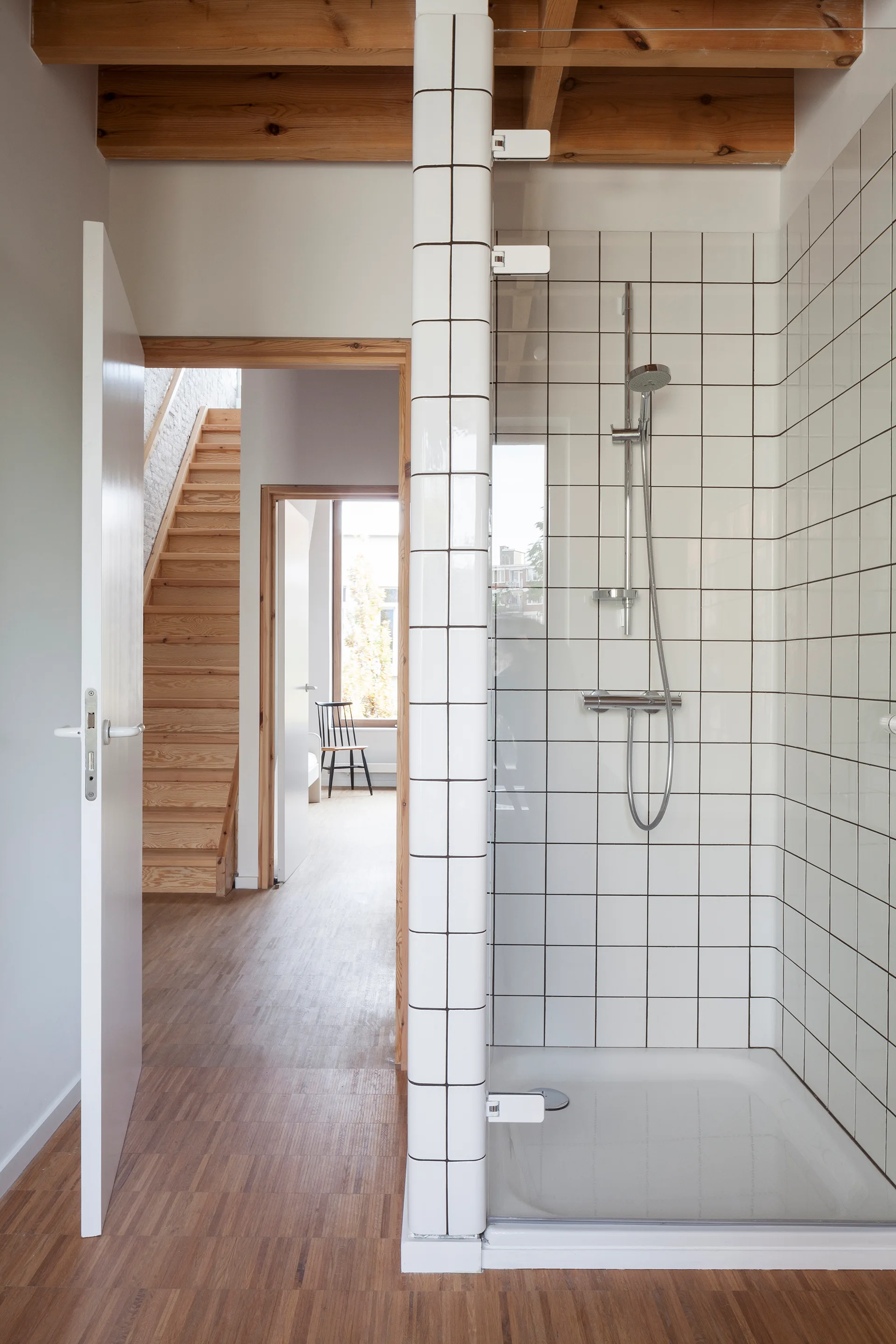 A modern bathroom featuring a white grid-tiled shower enclosure and exposed wooden ceiling beams with a view into a hallway.