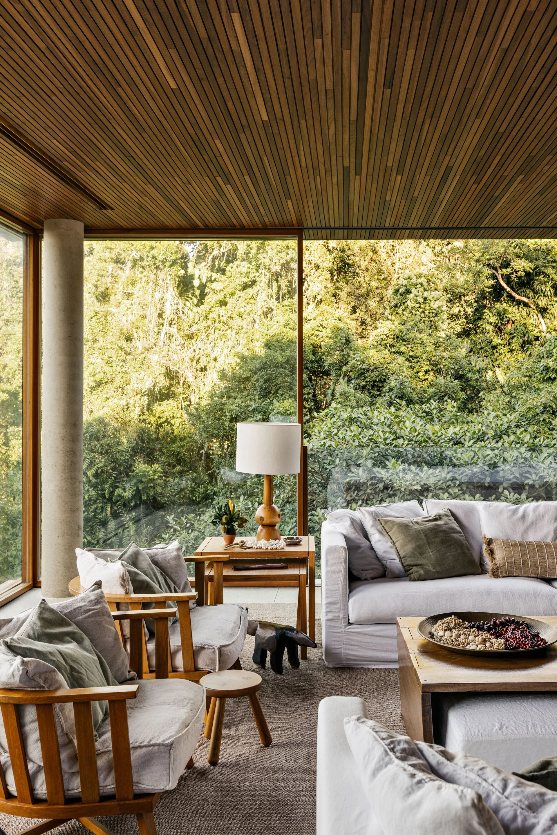 An organic modern living room with a slatted wood ceiling, neutral armchairs, and floor-to-ceiling windows displaying a lush forest backdrop.