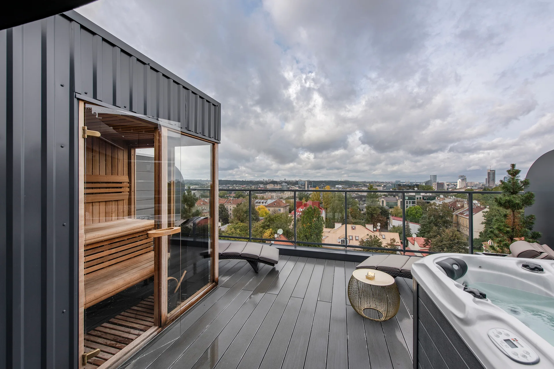 A grey composite rooftop deck features a glass-front wooden sauna, a white hot tub, and lounge chairs overlooking a city skyline.