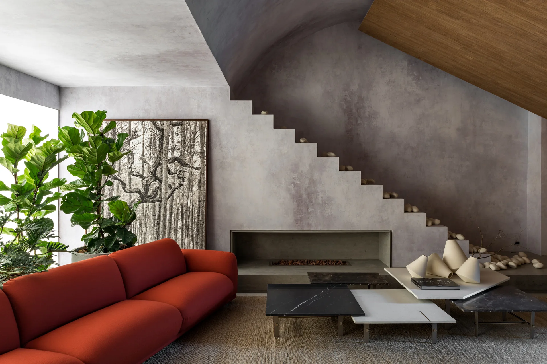 A modern living room features a bright red sofa, nested stone coffee tables, and a concrete staircase structure with a built-in fireplace.