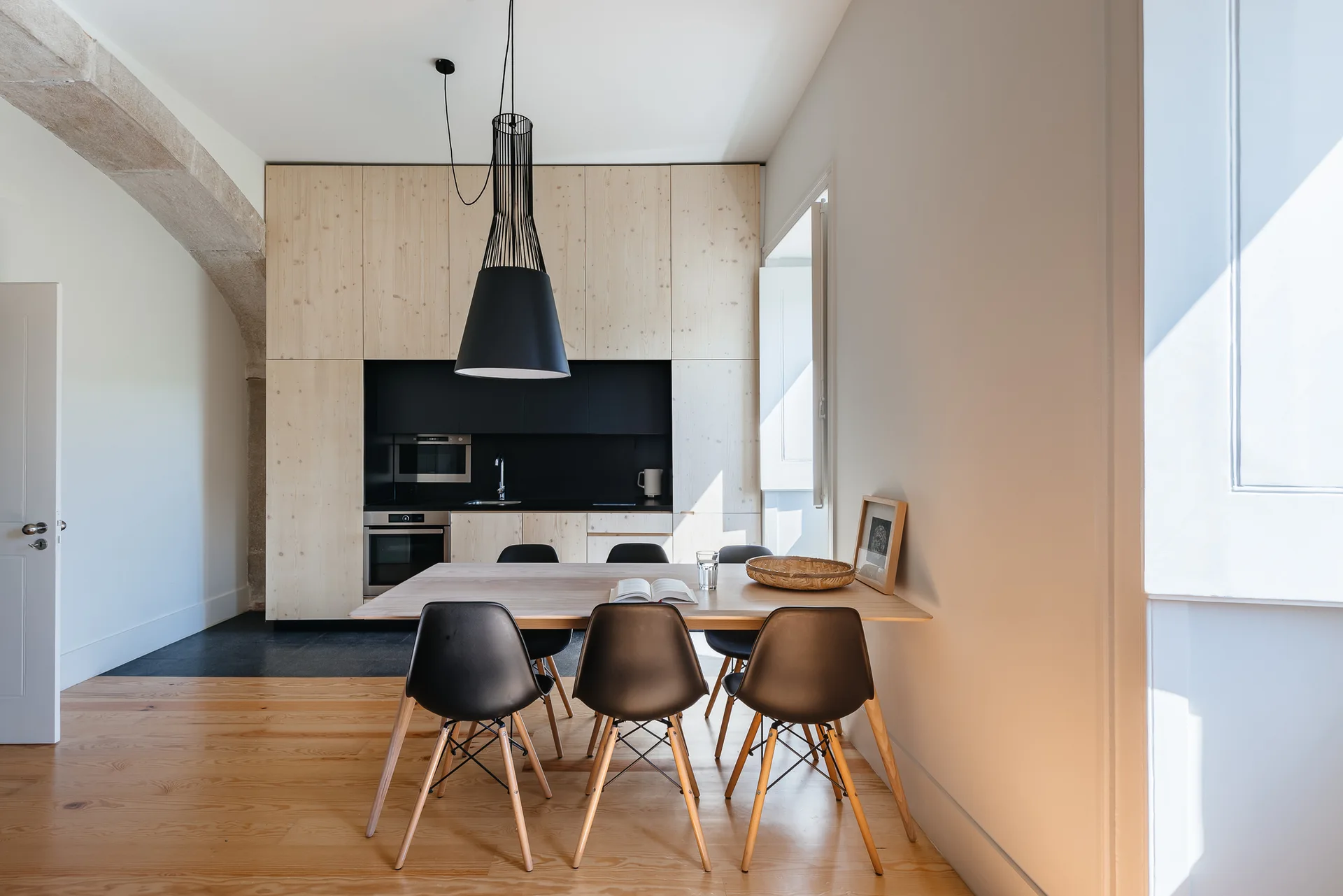 A minimalist kitchen with light wood cabinetry and a black recessed alcove next to a light wood dining table with black chairs.