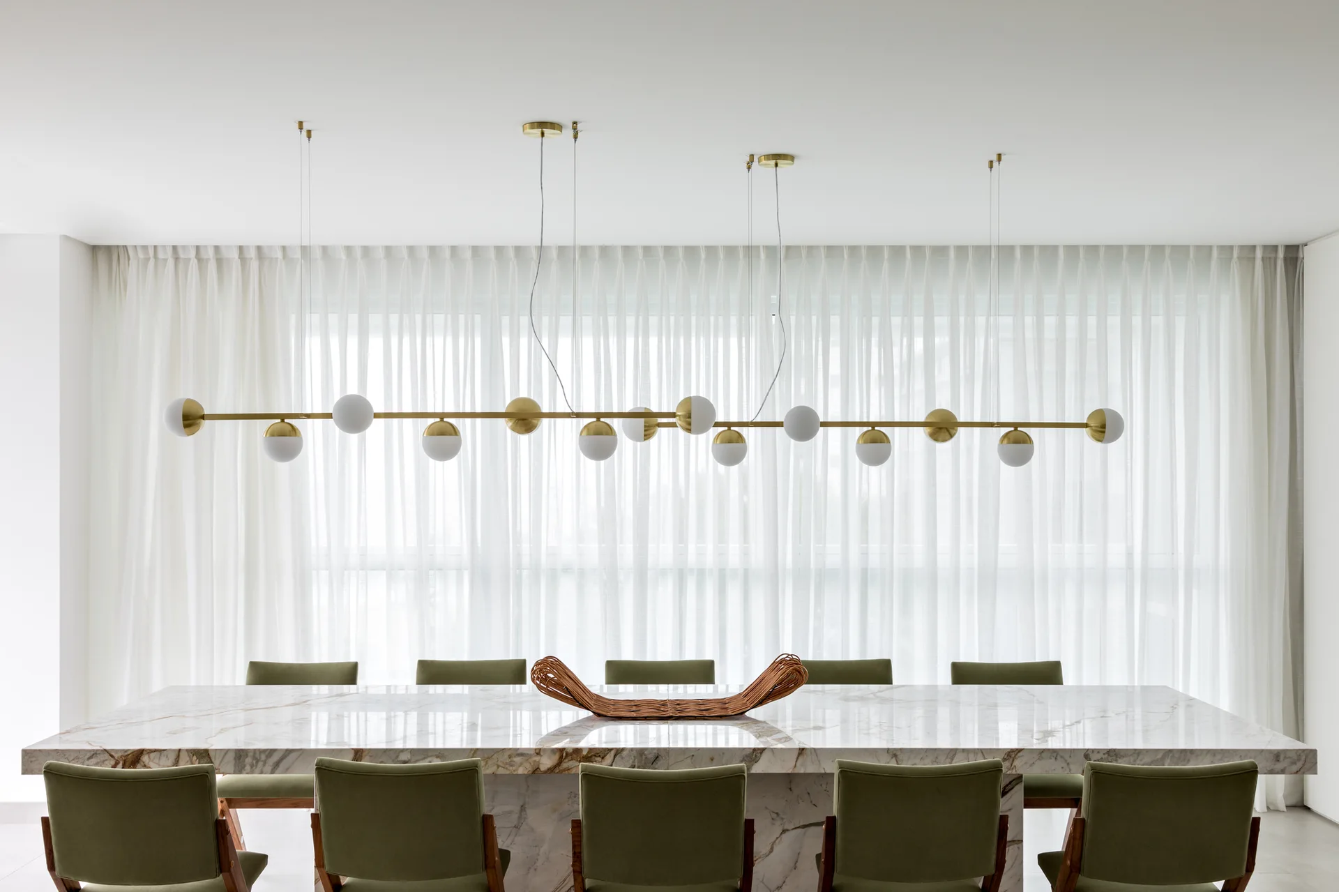 A long marble dining table is surrounded by olive green chairs under a linear brass pendant light with globe bulbs.