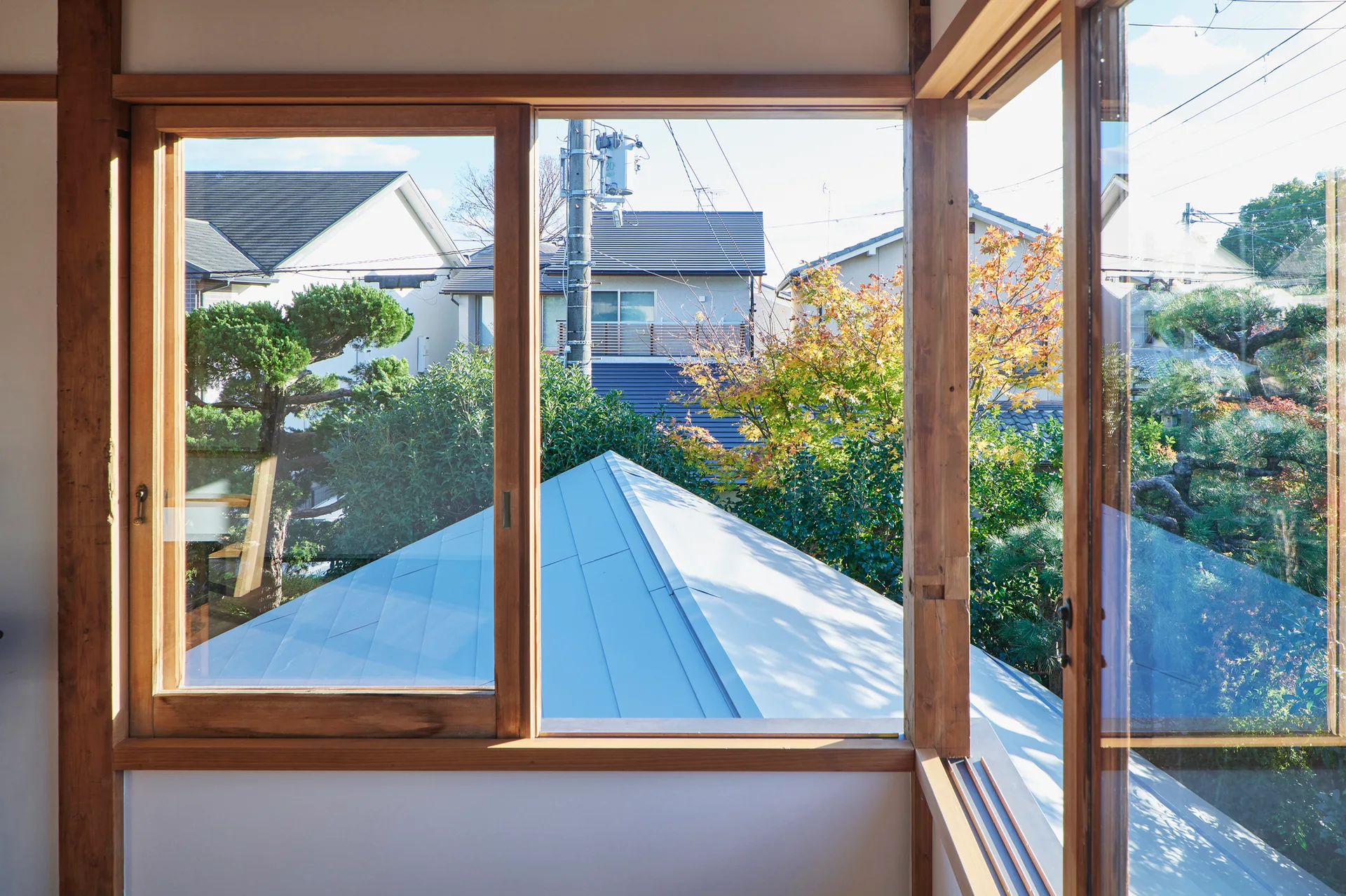 A view from inside a room looking through large natural wood windows out to neighboring houses and green trees.