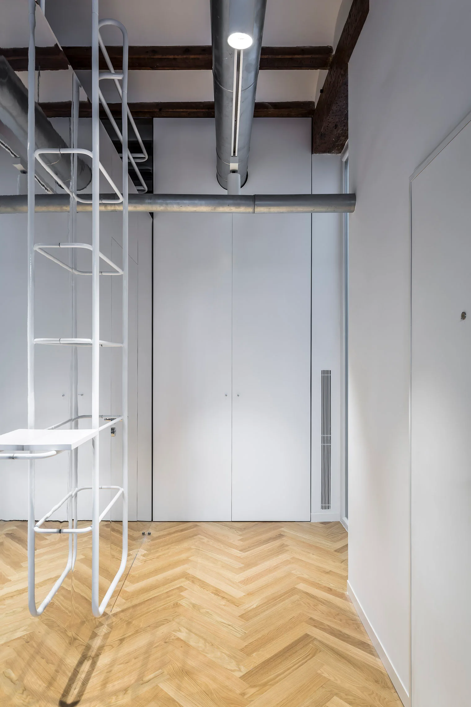 An industrial hallway featuring white built-in storage cabinets, a white metal ladder, herringbone wood floors, and exposed overhead metal ducts.