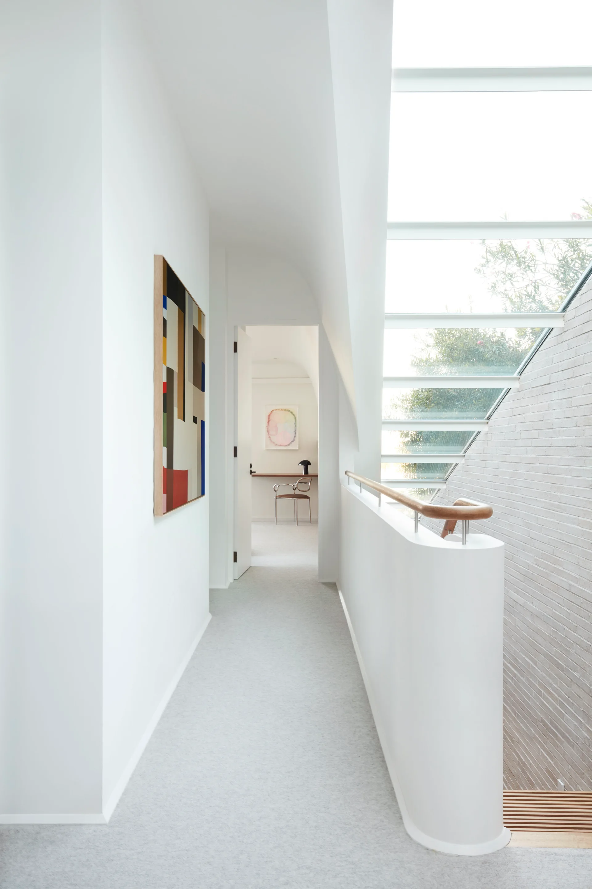 A bright white hallway features a large sloped skylight over a staircase, gray carpeted floors, and abstract framed art on the wall.
