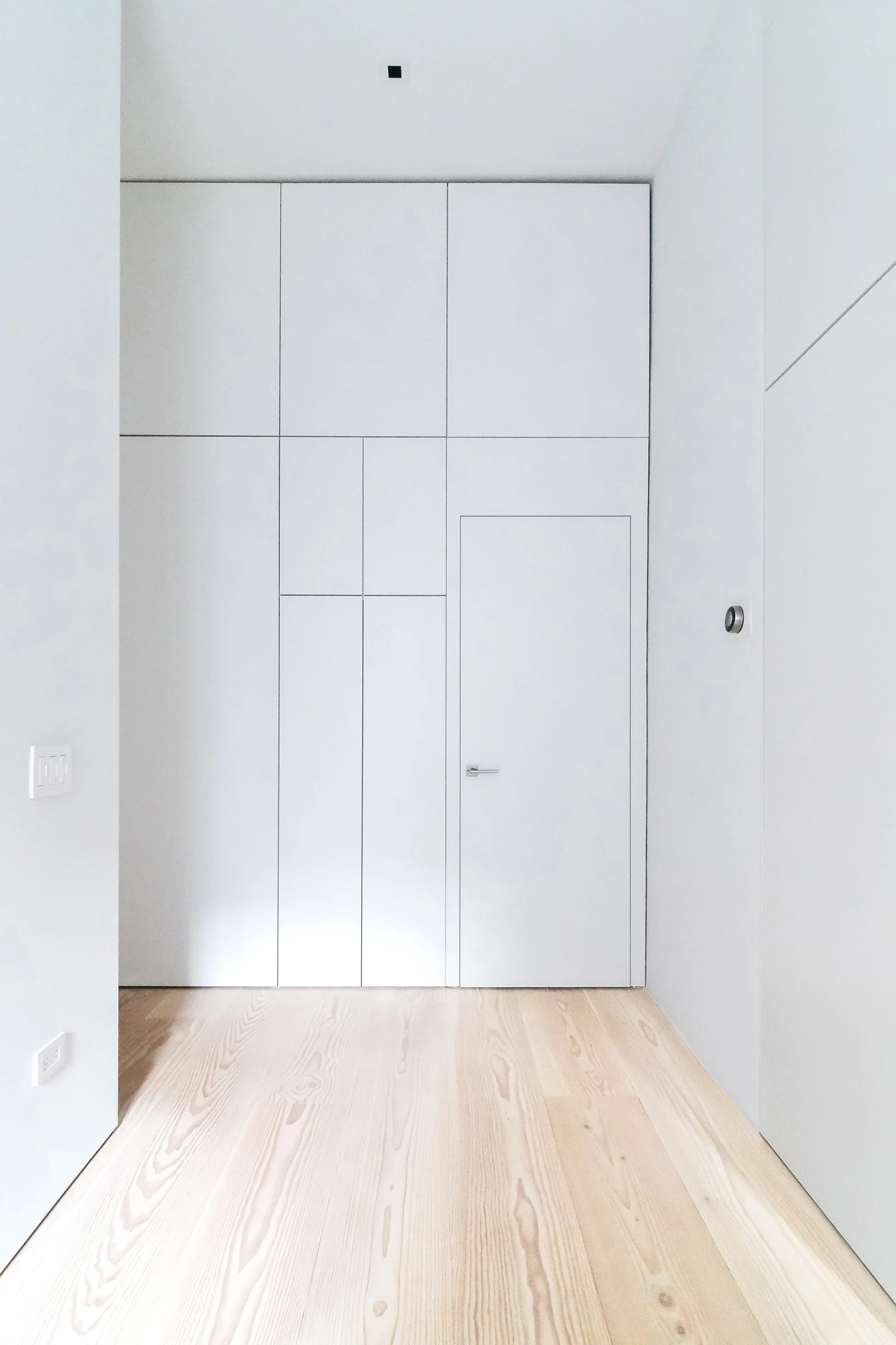 A minimalist hallway features a white floor-to-ceiling storage wall with an integrated flush door and light wood plank flooring.