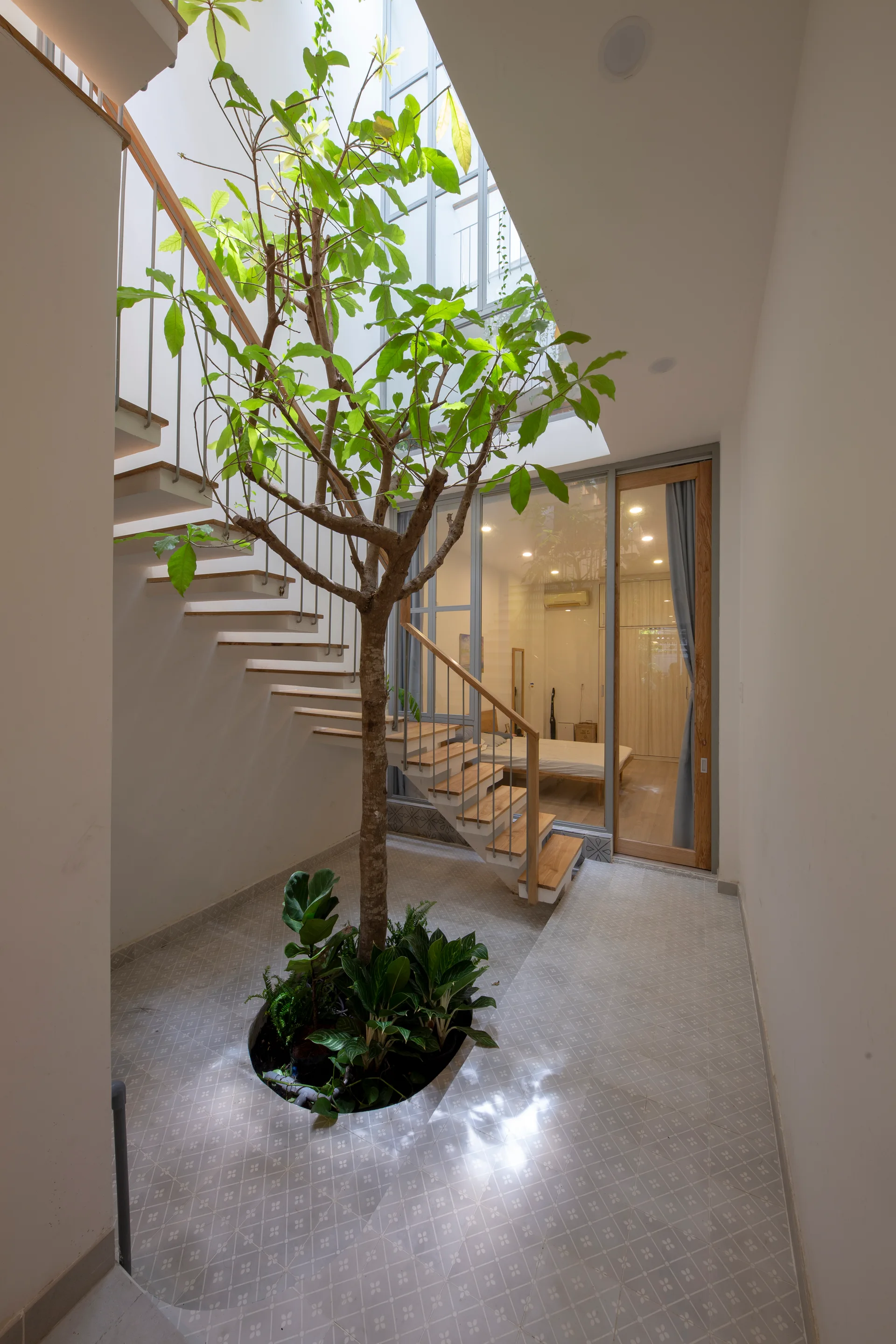 A wooden open-riser staircase wraps around a tall indoor tree planted in a circular floor cutout beneath a bright skylight.