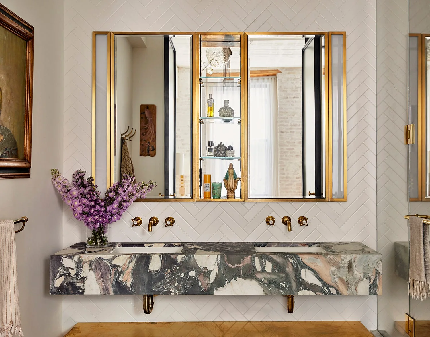 A wall-mounted marble vanity with dual sinks sits against a white herringbone tile wall beneath brass-trimmed mirrors and wall-mounted gold faucets.