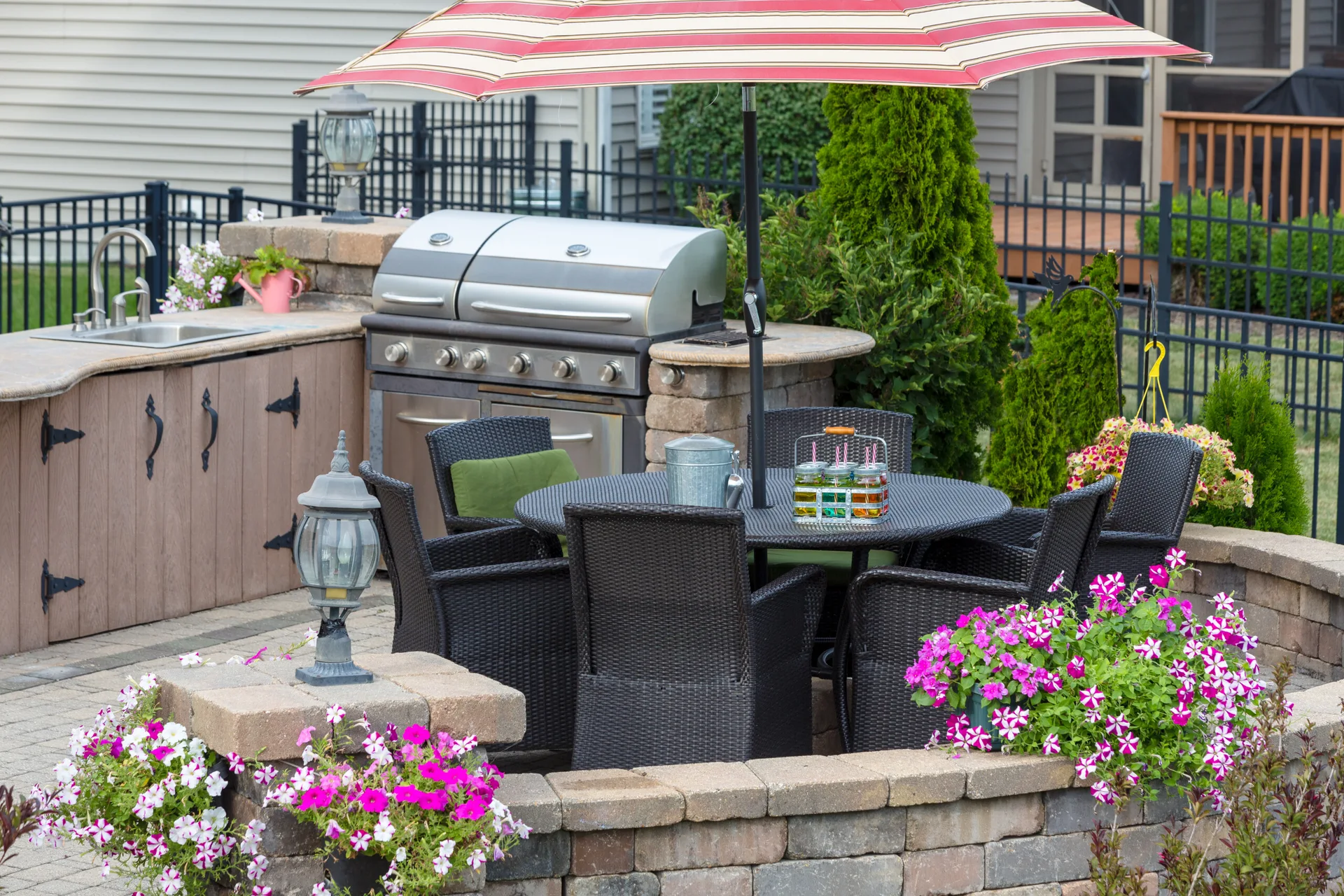 A backyard stone patio features a built-in outdoor kitchen, a dark wicker dining set under a striped umbrella, and low stone retaining walls with pink flowers.