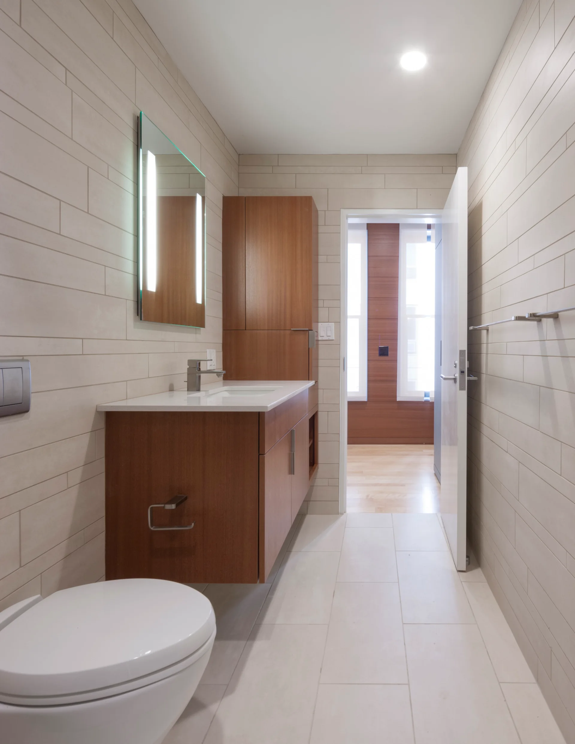 A modern minimalist bathroom featuring a wall-mounted wood vanity, a backlit mirror, and beige rectangular wall tiles.