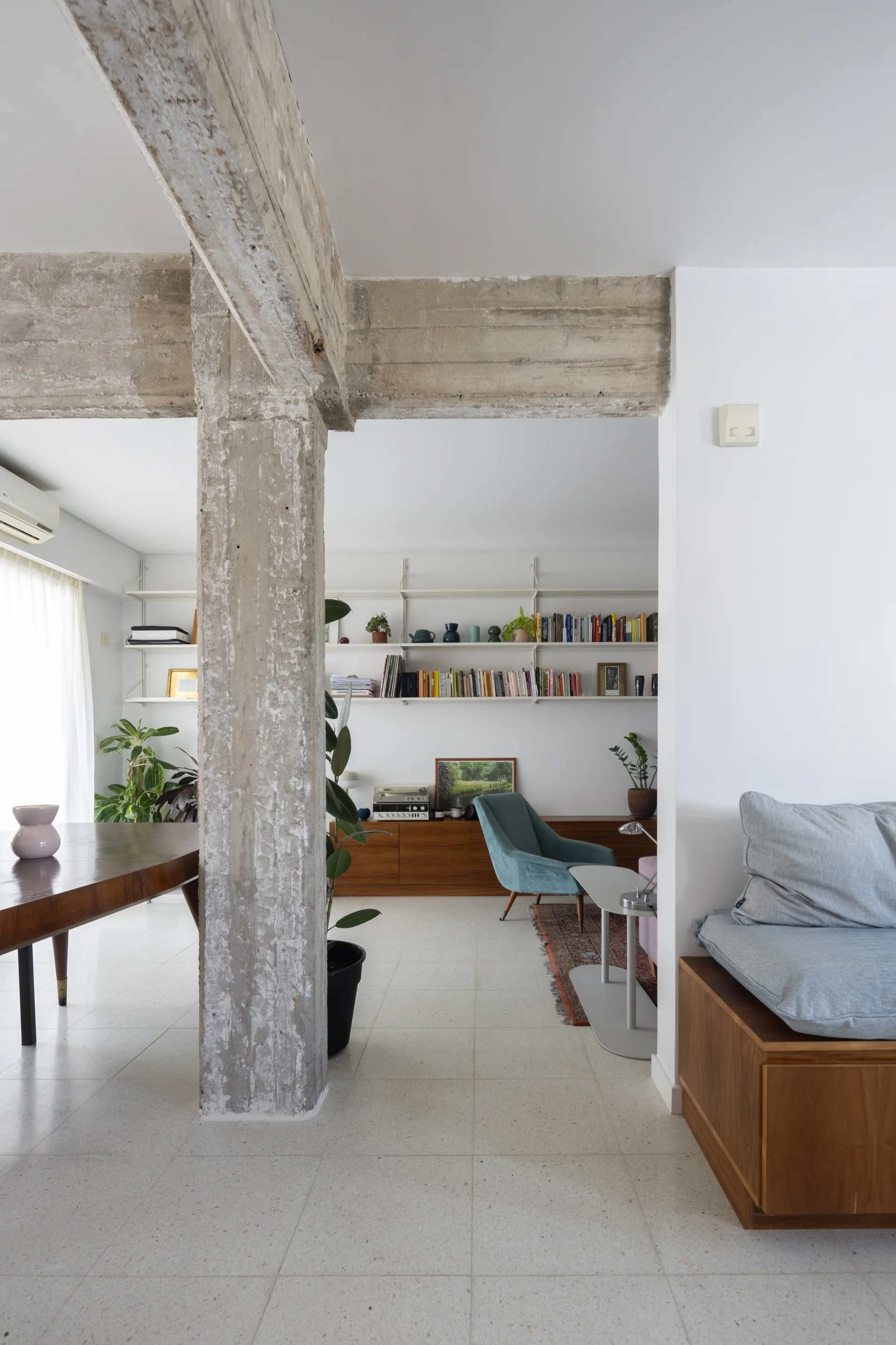An open-plan living space features an exposed concrete structural column, low wooden cabinets, white wall shelving, and a blue armchair.