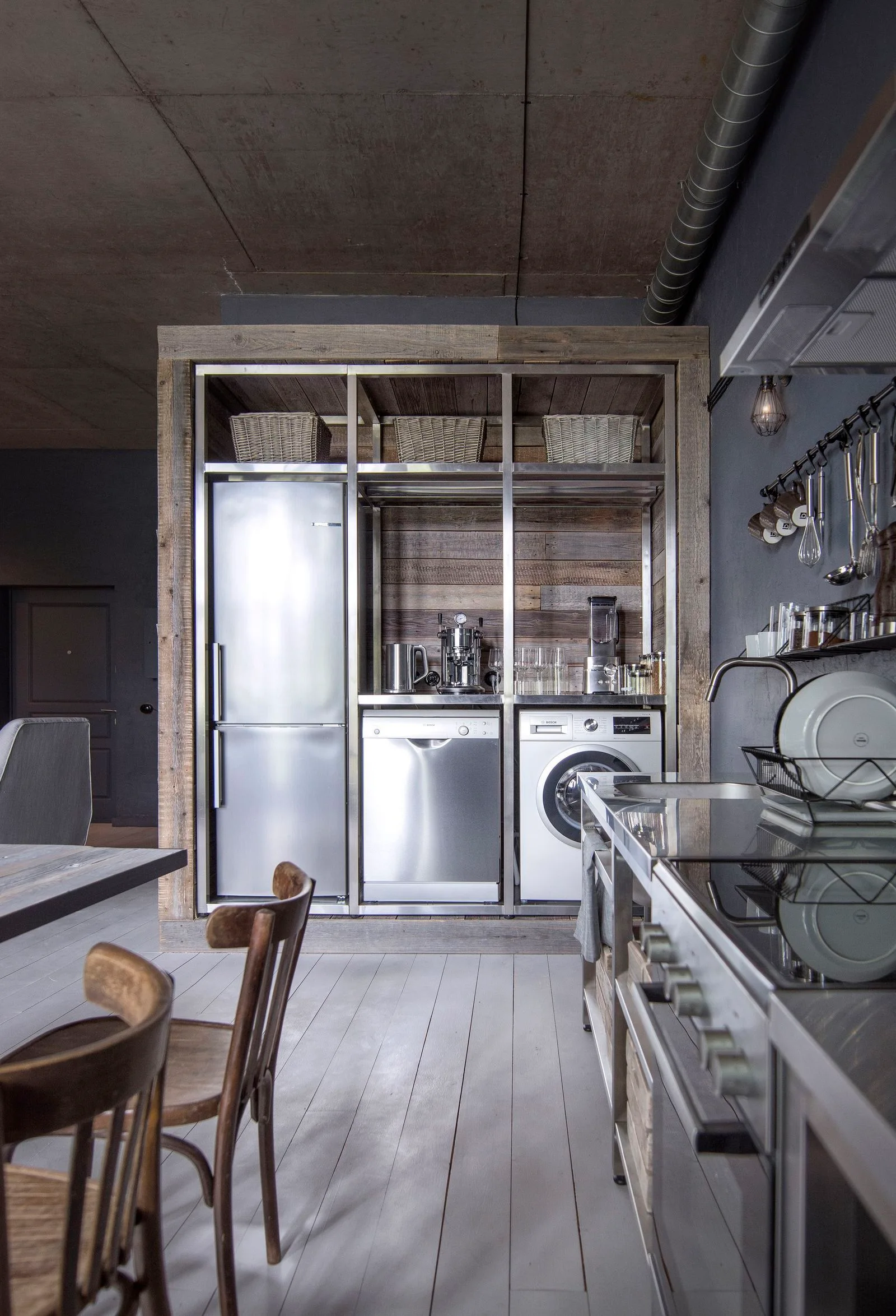 An industrial kitchen features a large custom unit of steel and reclaimed wood housing a stainless steel refrigerator, dishwasher, and washing machine.
